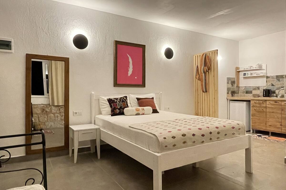 A bedroom with a bed, bedside tables, a lamp, a mirror, and a small kitchenette.