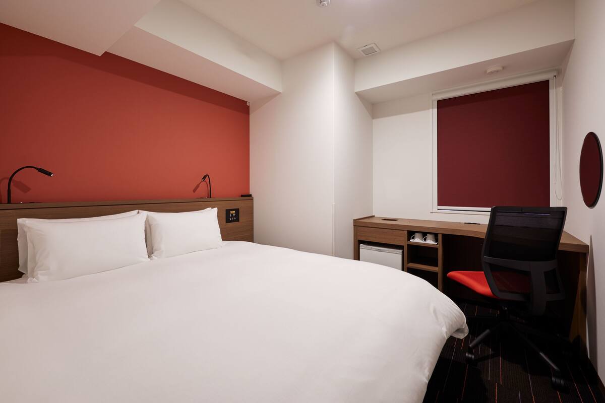 A hotel room with a large bed, a desk with a chair, a small table, and a window with blinds.