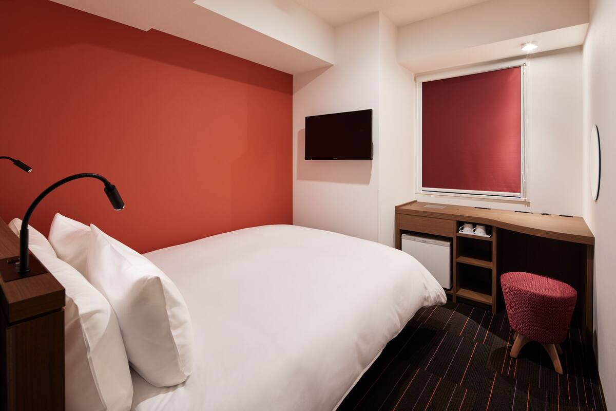 A hotel room with a bed, a desk, a chair, a TV, and a window with a blind.