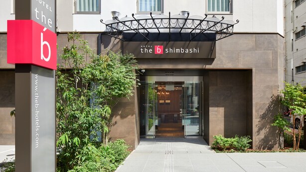 L’entrée de l’hôtel b Shimbashi présente un design moderne, avec une porte en verre et un auvent métallique au-dessus de l’entrée.