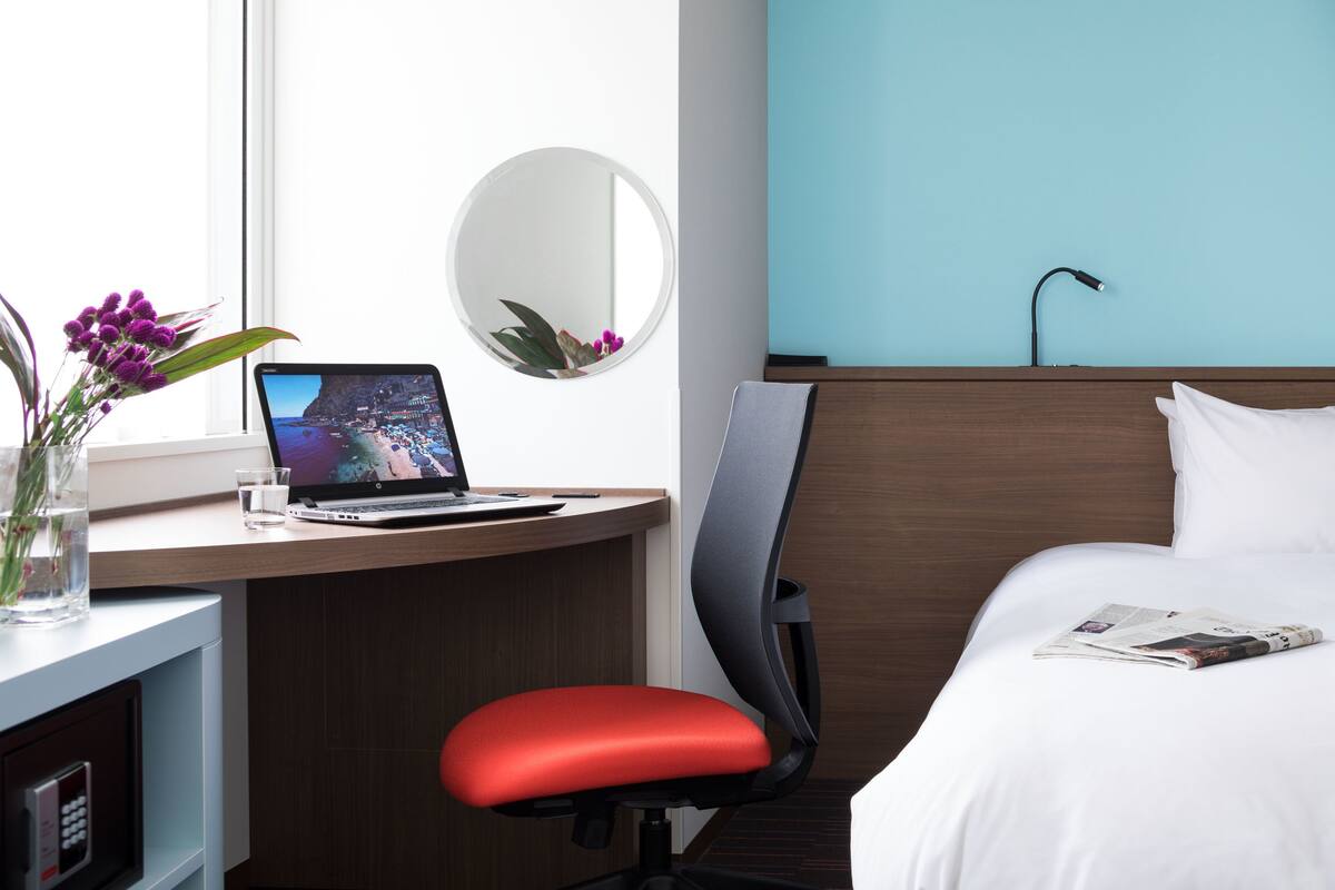 A hotel room with a desk, laptop, chair, bed, and a round mirror.