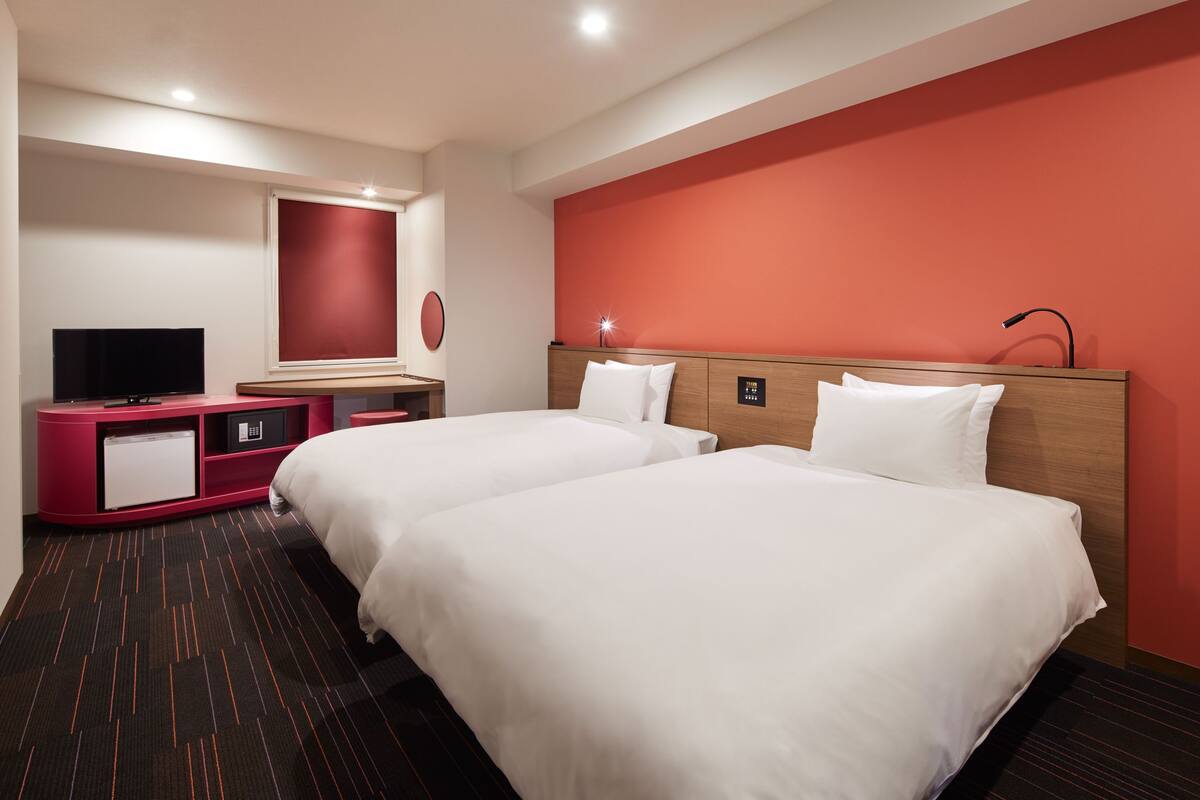 A hotel room with two beds, a TV, a desk, and a red accent wall.