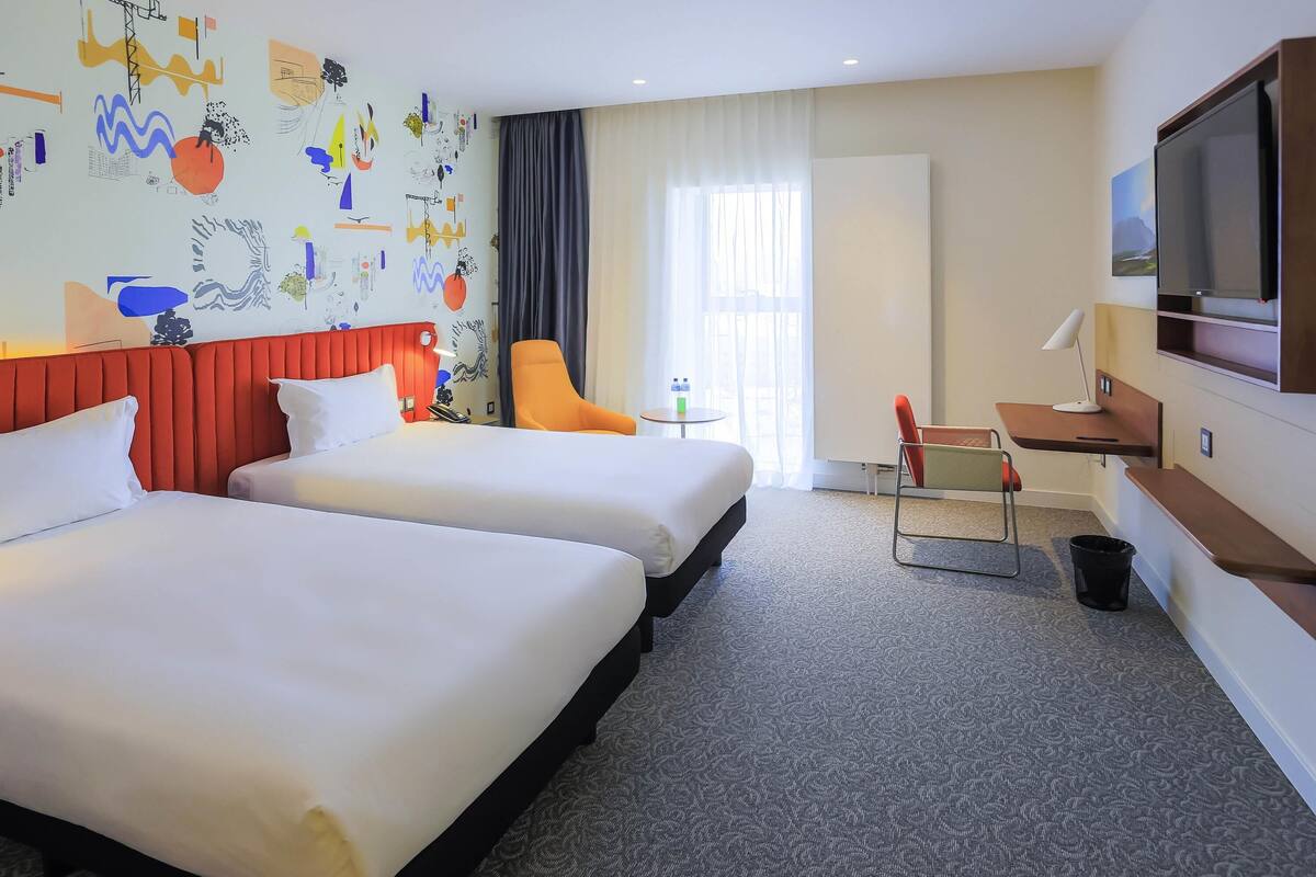 A hotel room with two beds, a desk, a TV, and a window with curtains.