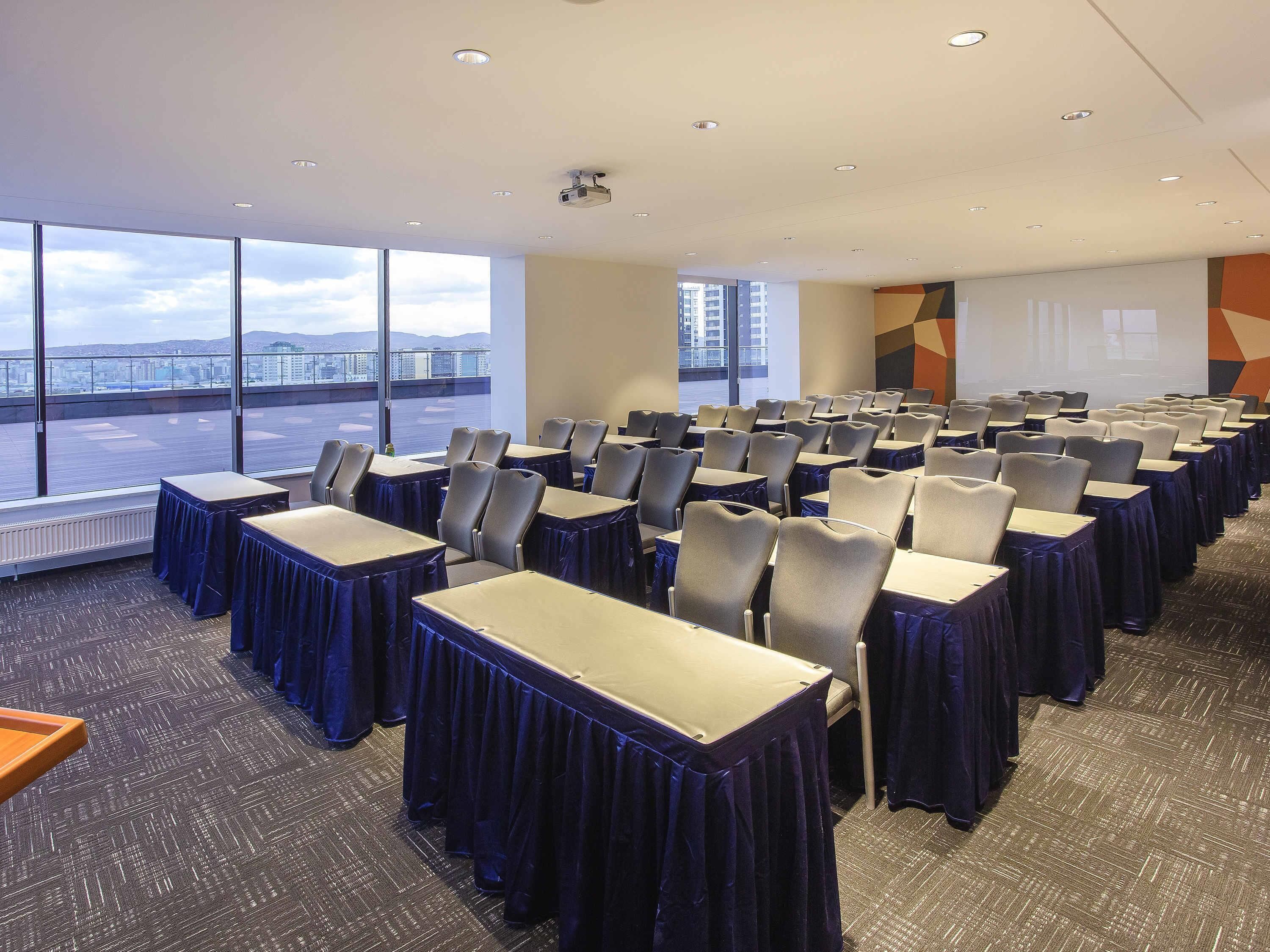 Un salón de conferencias con filas de mesas y sillas, ventanas amplias que ofrecen vistas a la ciudad y una pantalla para proyector.