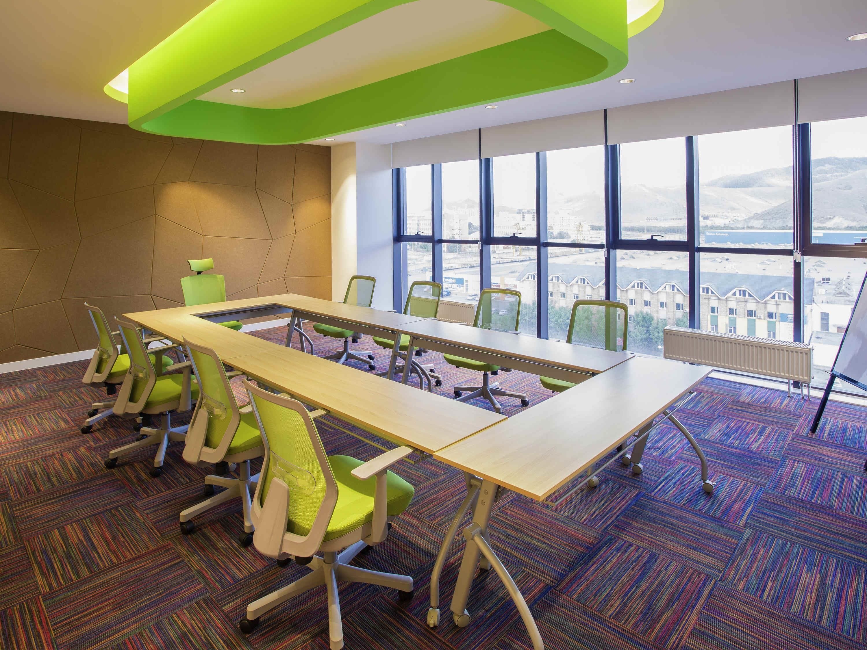Een congreszaal met een lange tafel, groene stoelen en grote ramen met uitzicht op de stad.