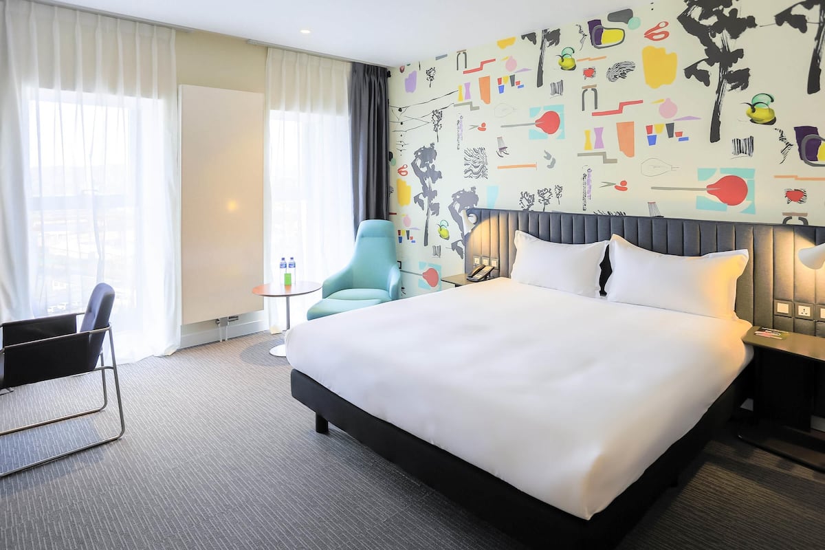 A hotel room with a large bed, a chair, a small table, and a wall with abstract art.