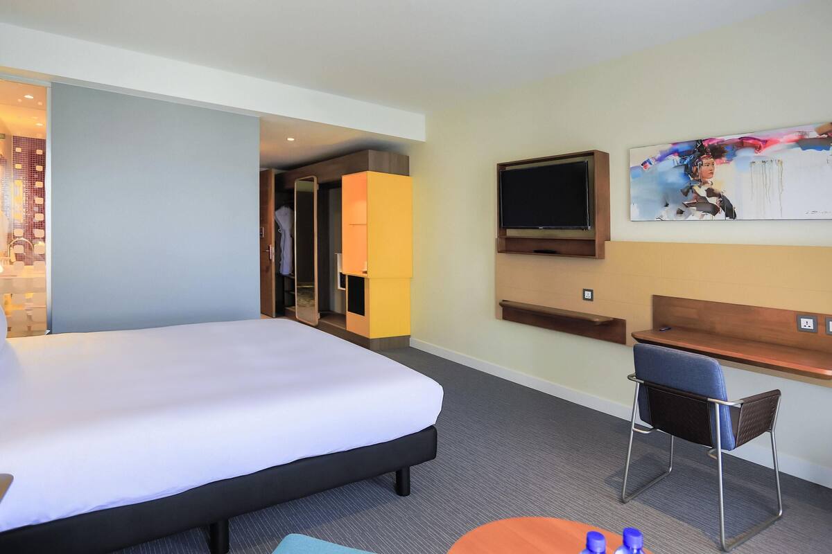 A hotel room with a bed, a desk, a chair, a TV, and a painting on the wall.