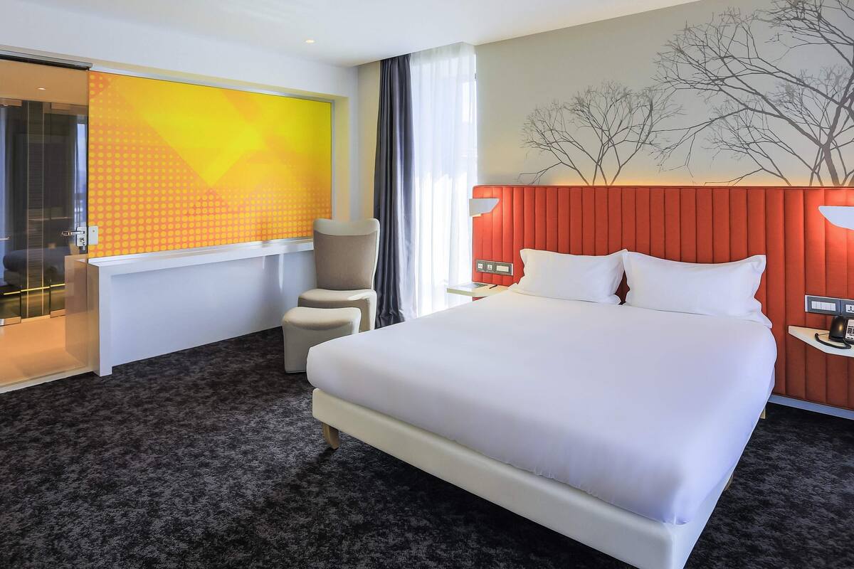 A hotel room with a large bed, a desk, a chair, a window with curtains, and a wall with a tree mural.