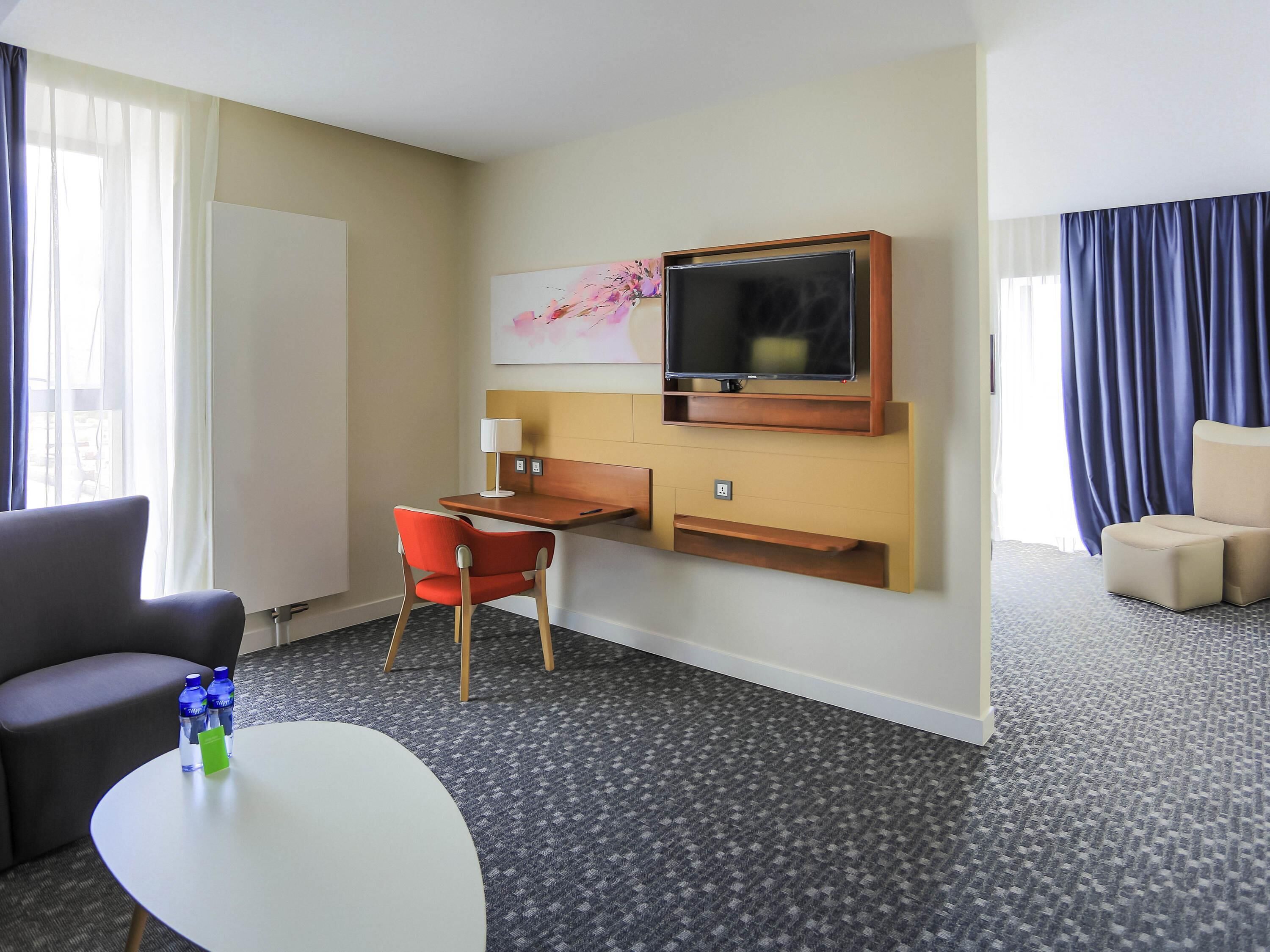 A hotel room with a desk, a TV mounted on the wall, a chair, a sofa, and a small table with a water bottle and a green card.