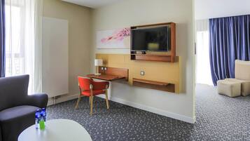 A hotel room with a desk, a TV mounted on the wall, a chair, a sofa, and a small table with a water bottle and a green card.
