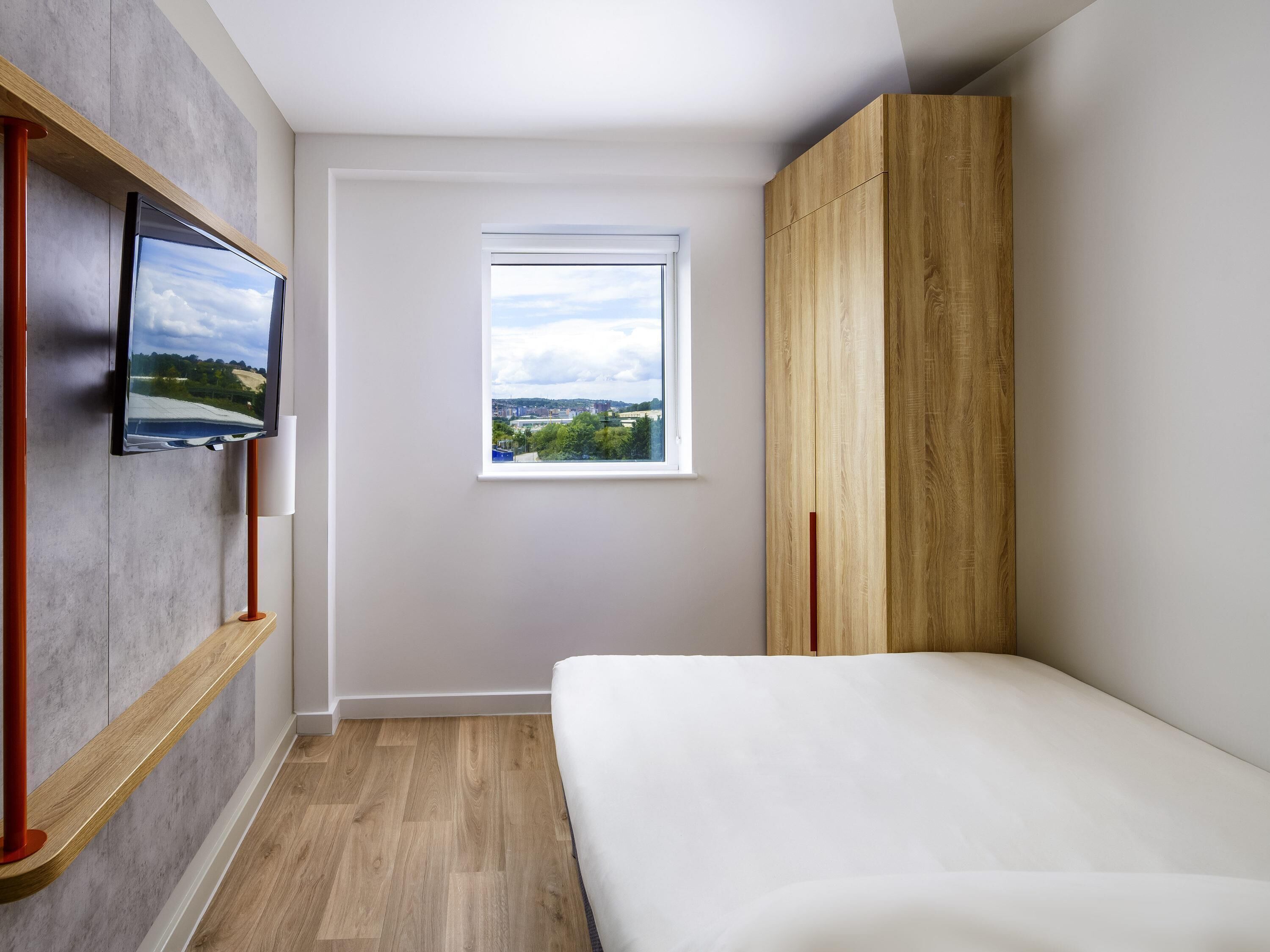 Ein Schlafzimmer mit einem Bett, einem an der Wand montierten Fernseher, einem Fenster mit Blick auf die Stadt und einem Holzschrank.