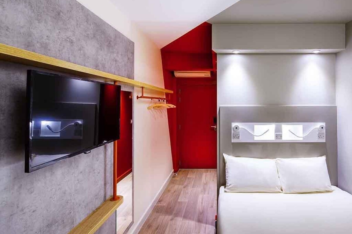 A hotel room with a flat-screen TV mounted on the wall, a bed with white bedding, and a red accent wall.