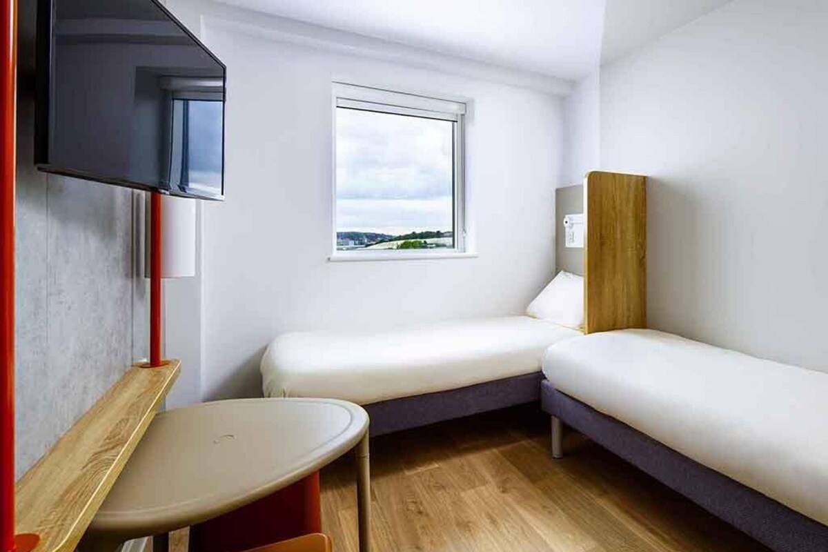 A small, modern room with two single beds, a wooden table, a TV mounted on the wall, and a window with a view.