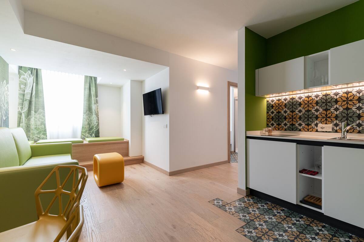 Una sala moderna con un sofá verde, un reposapiés amarillo y una mesa de centro de madera. Un televisor de pantalla plana está montado en la pared, y se ve una zona de cocina con armarios blancos y un salpicadero con diseño.