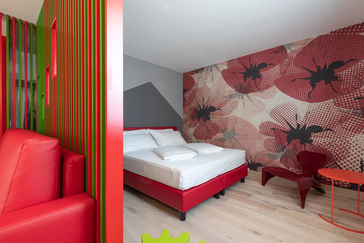 Una habitación de hotel moderna con una cama, una silla y una mesa rojas, y una pared a rayas de colores.