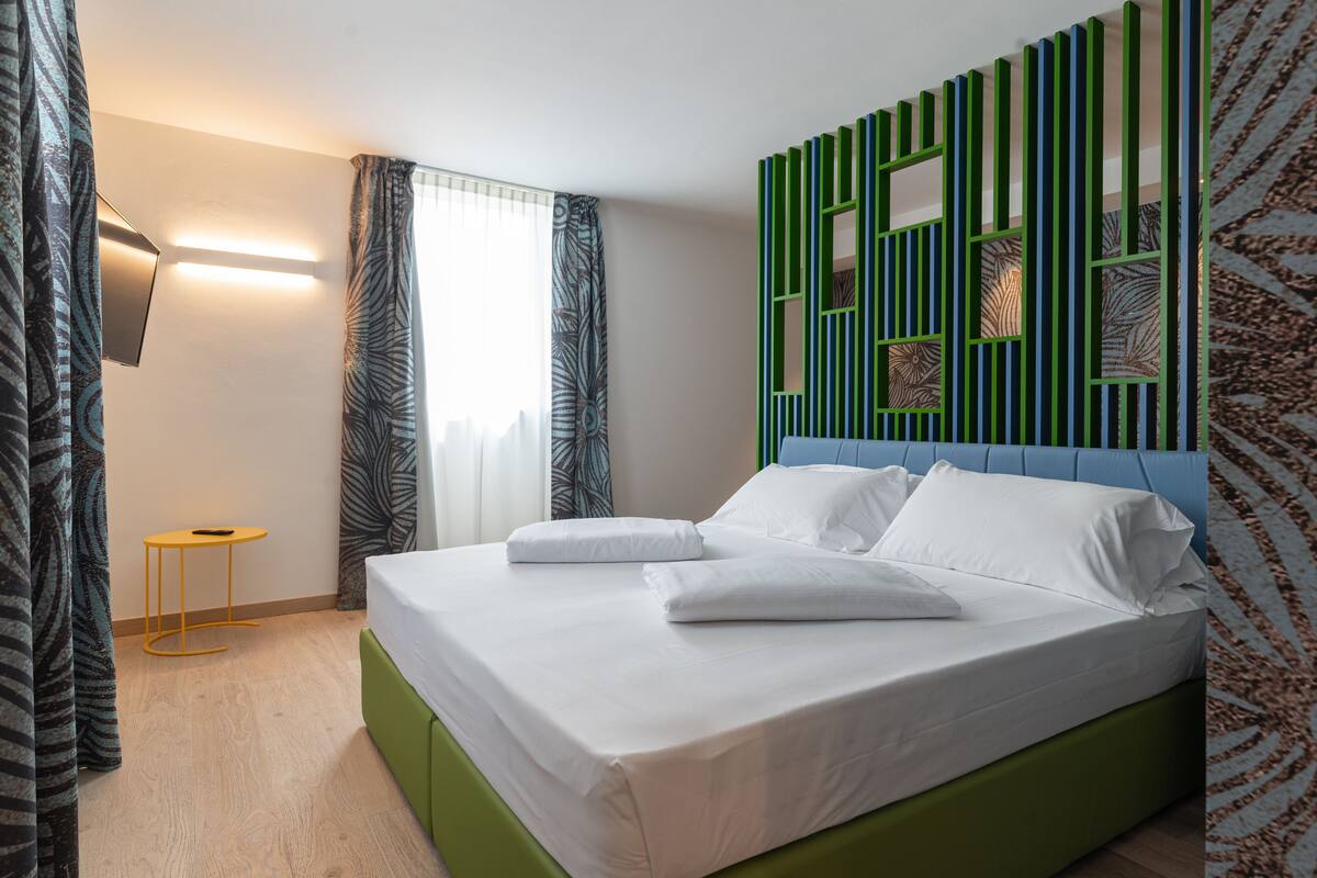 Una cama bien tendida con sábanas blancas en una habitación moderna con una pared de acento verde, una mesita auxiliar y cortinas estampadas.