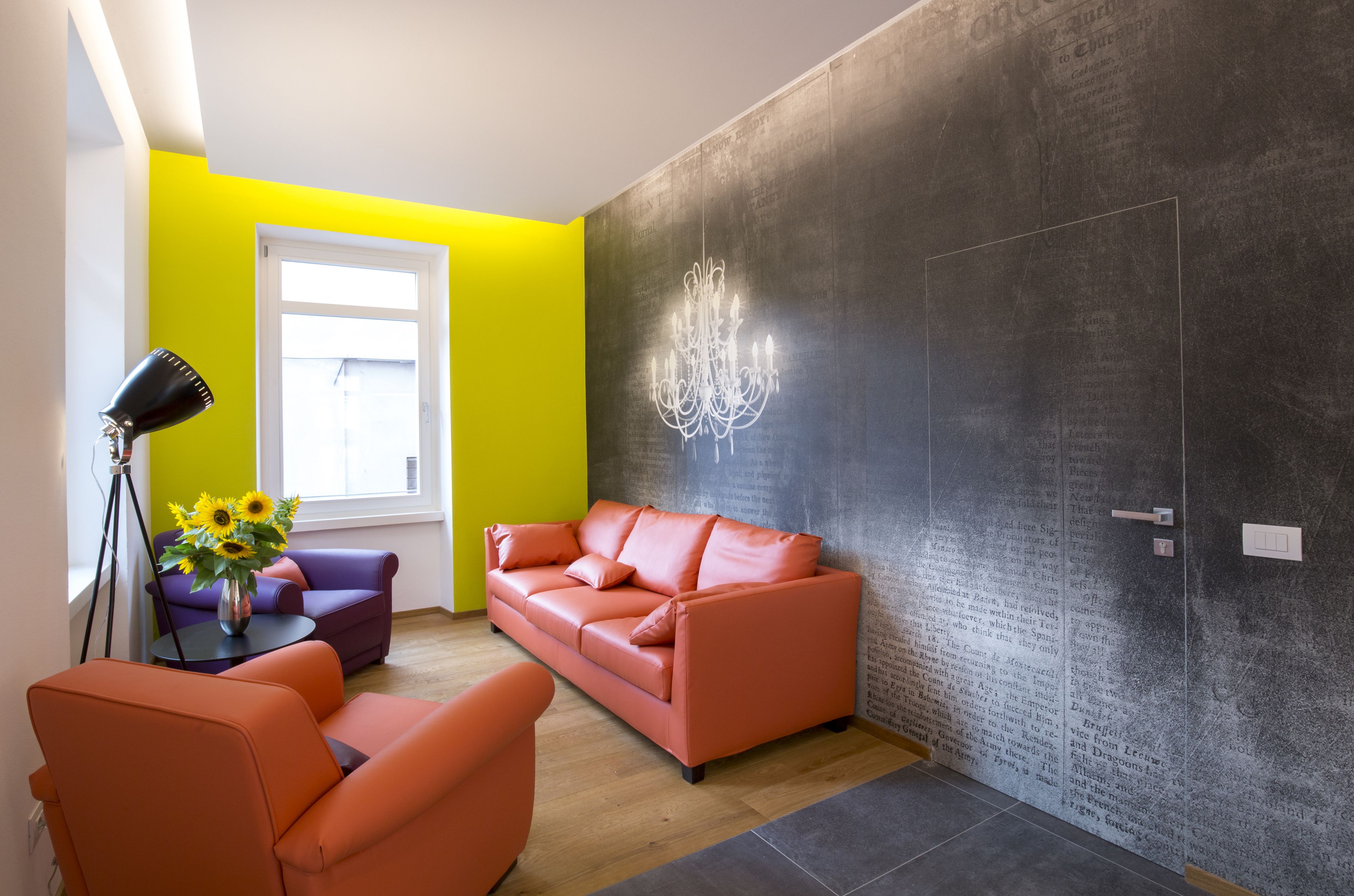 A modern living room with a bright yellow accent wall, a leather sofa, a small round table with a lamp, and a wall-mounted chandelier.