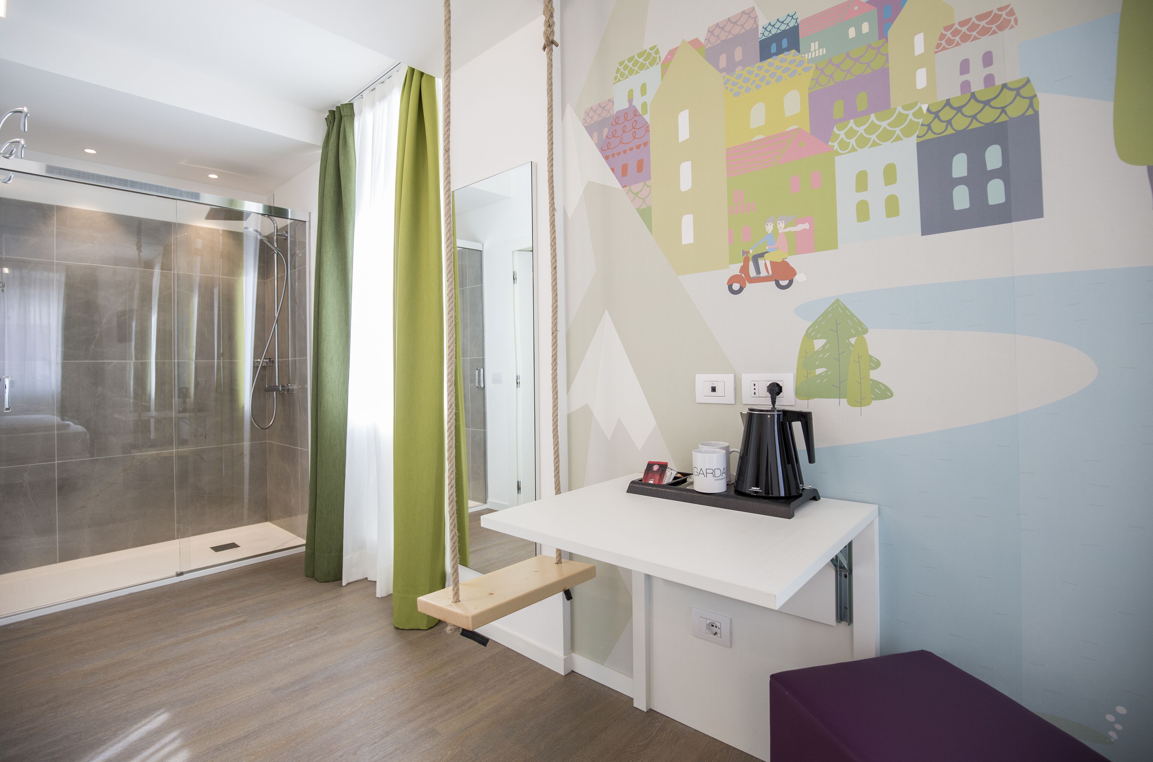 A modern bathroom with a glass shower enclosure, a green curtain, a white sink with a coffee maker, and a colorful mural on the wall.