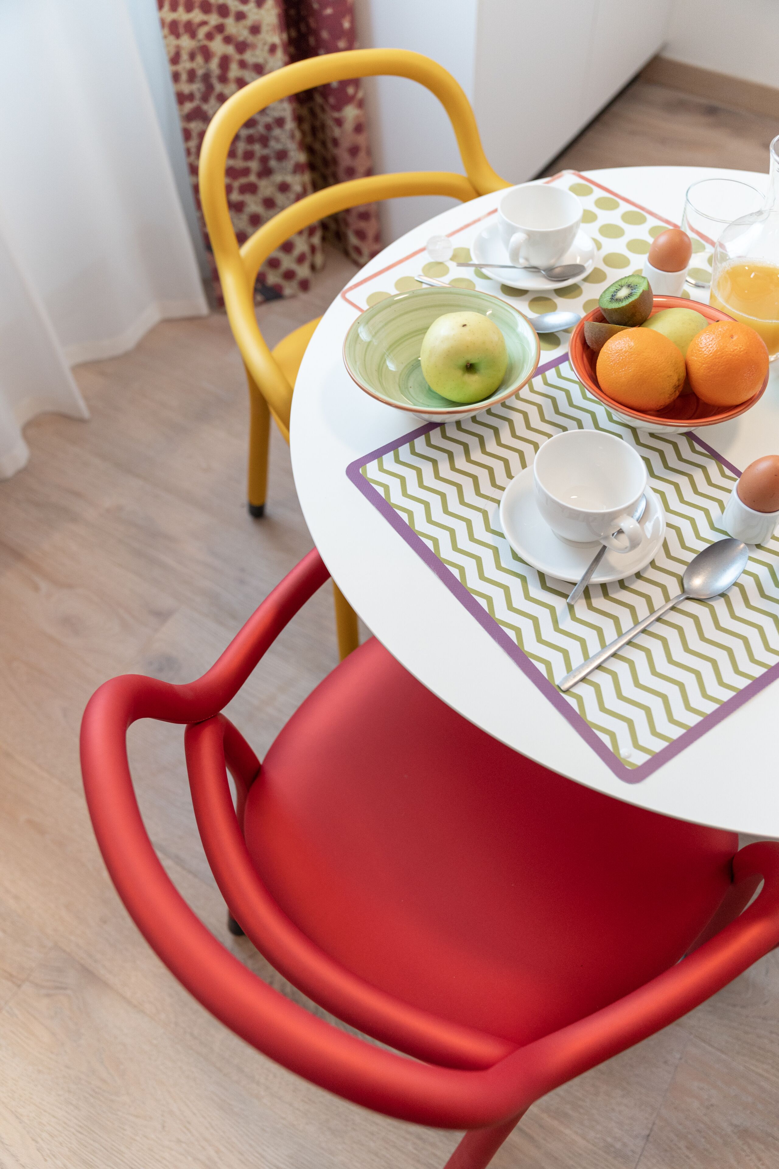 A dining table with a white and green patterned tablecloth, a red chair, and a yellow chair. The table is set with a glass, a bowl of fruit, and a cup.