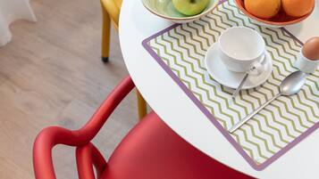 Una mesa de comedor con mantel blanco y verde estampado, una silla roja y una amarilla. La mesa está puesta con un vaso, un bol de fruta y una taza.