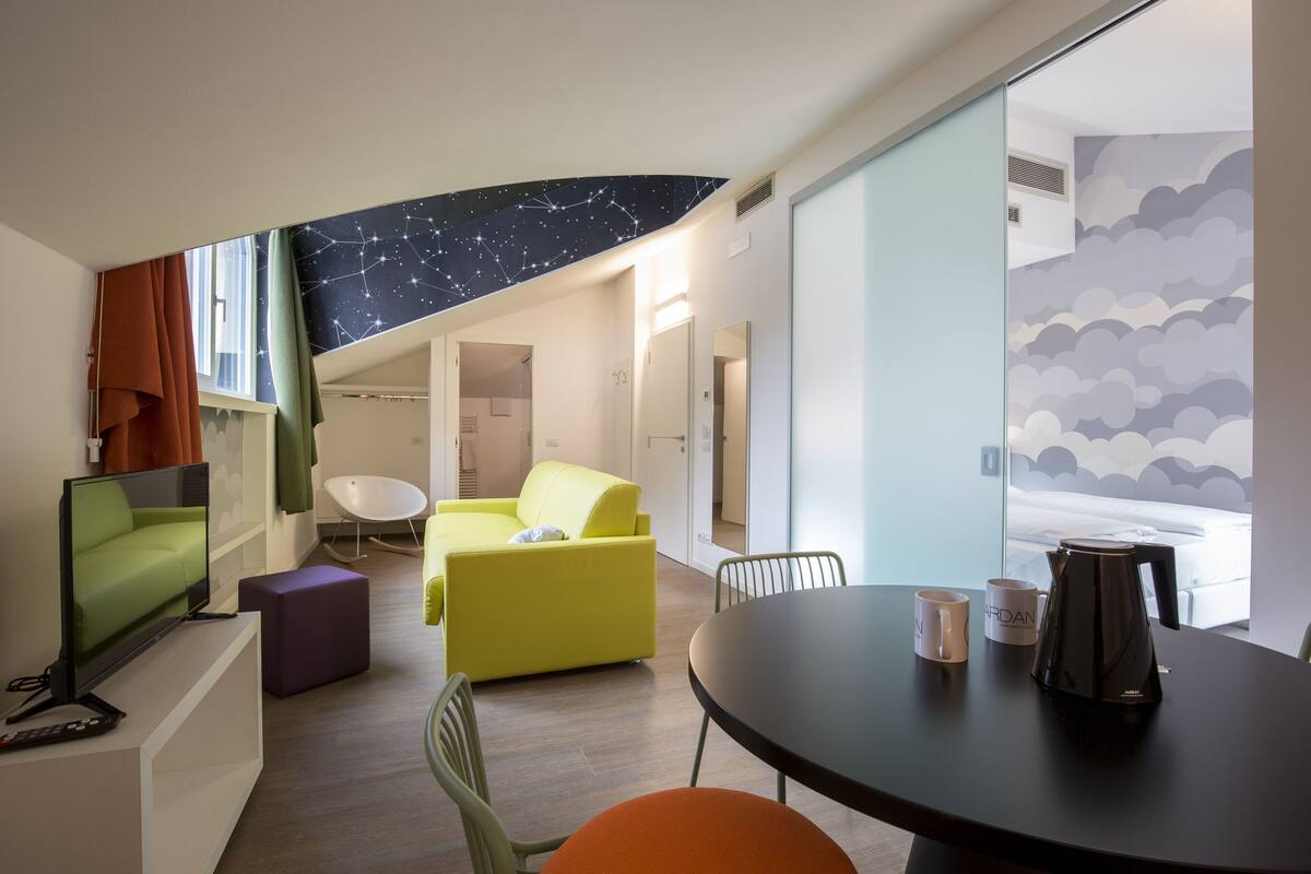 Una habitación de hotel moderna con techo estrellado, un sofá amarillo, un sillón verde, un reposapiés púrpura, una mesa redonda con dos tazas y una cafetera.