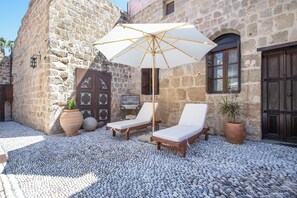 Un edificio de piedra con patio, dos sillones, una sombrilla grande y plantas en macetas.