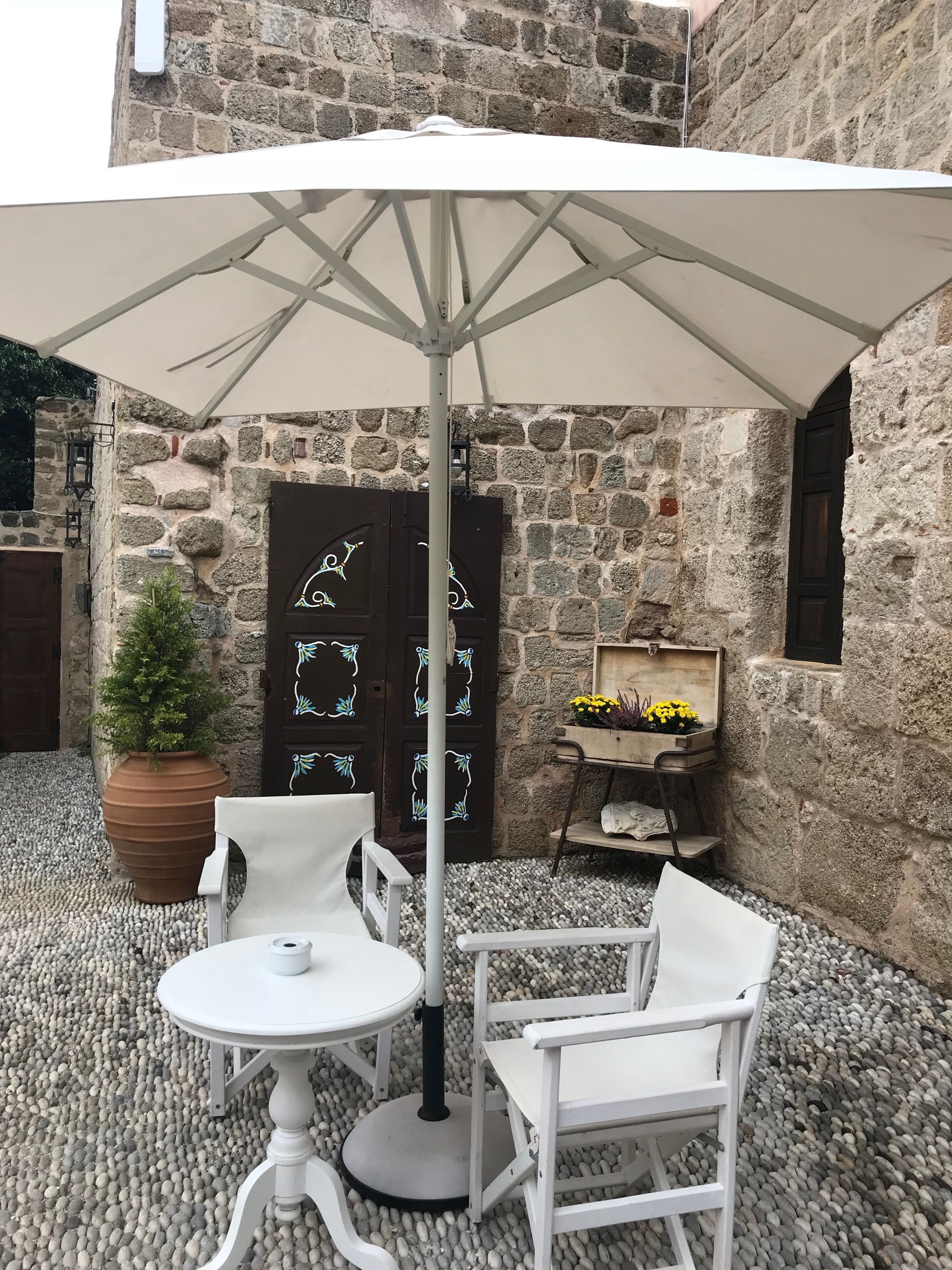 An outdoor seating area with a table and chairs under a white umbrella, set against a stone wall with a wooden door and a potted plant.