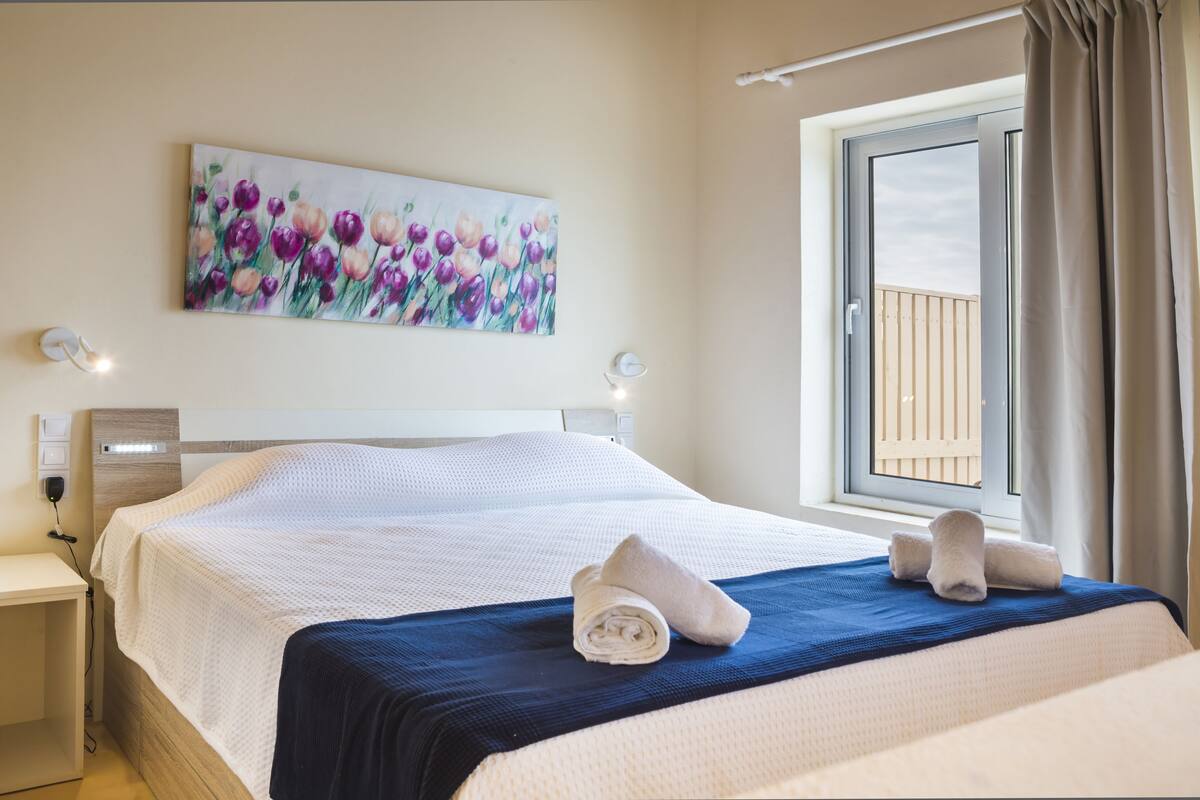 Un lit bien fait, recouvert d’une couvre-lit bleue et blanche, un tableau de fleurs accroché au mur et une fenêtre avec des rideaux.