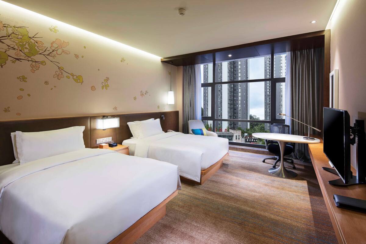 A hotel room with two beds, a desk, a chair, a TV, and a large window with a city view.
