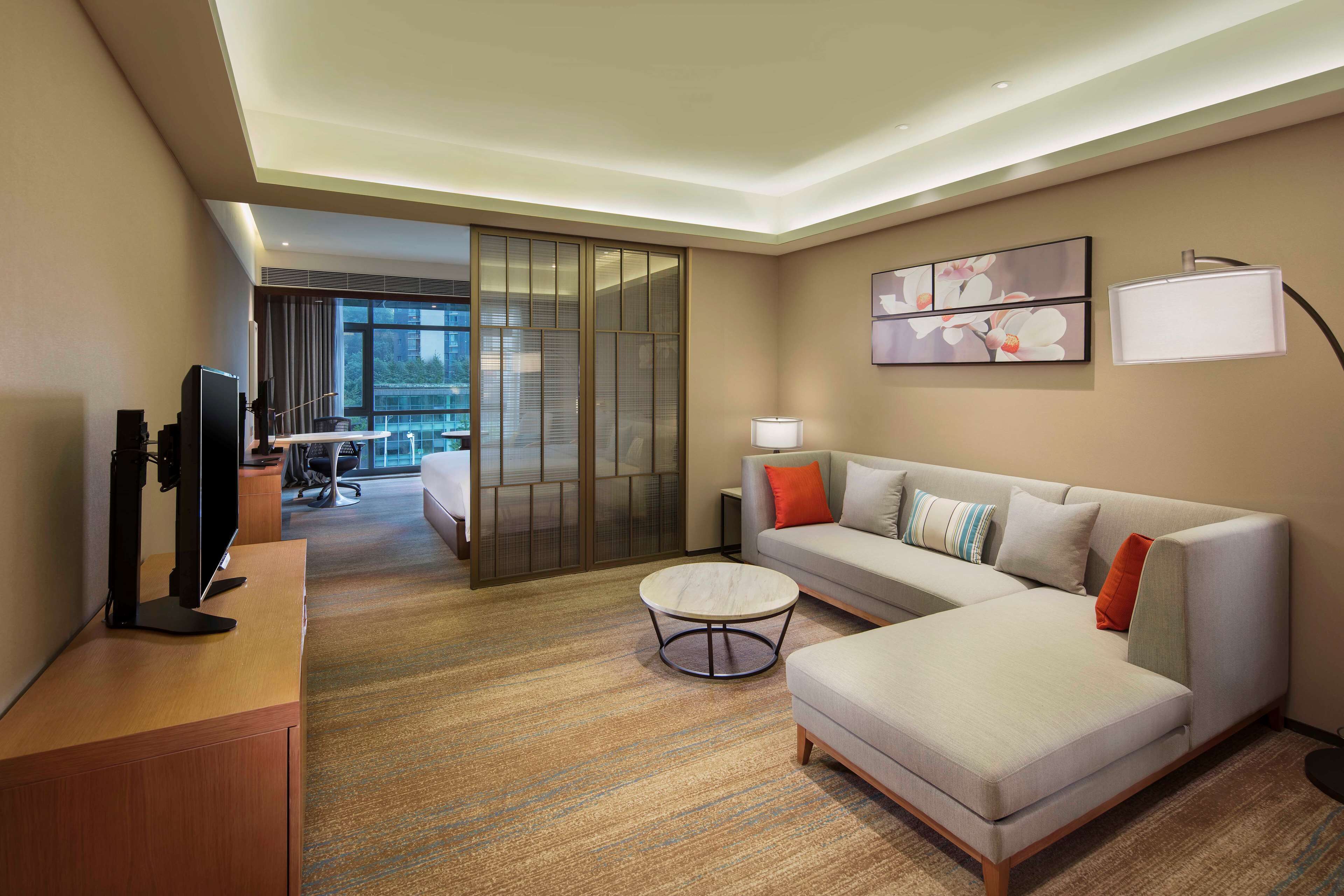 A modern hotel room with a sofa, a coffee table, a TV, and a sliding door leading to a balcony.