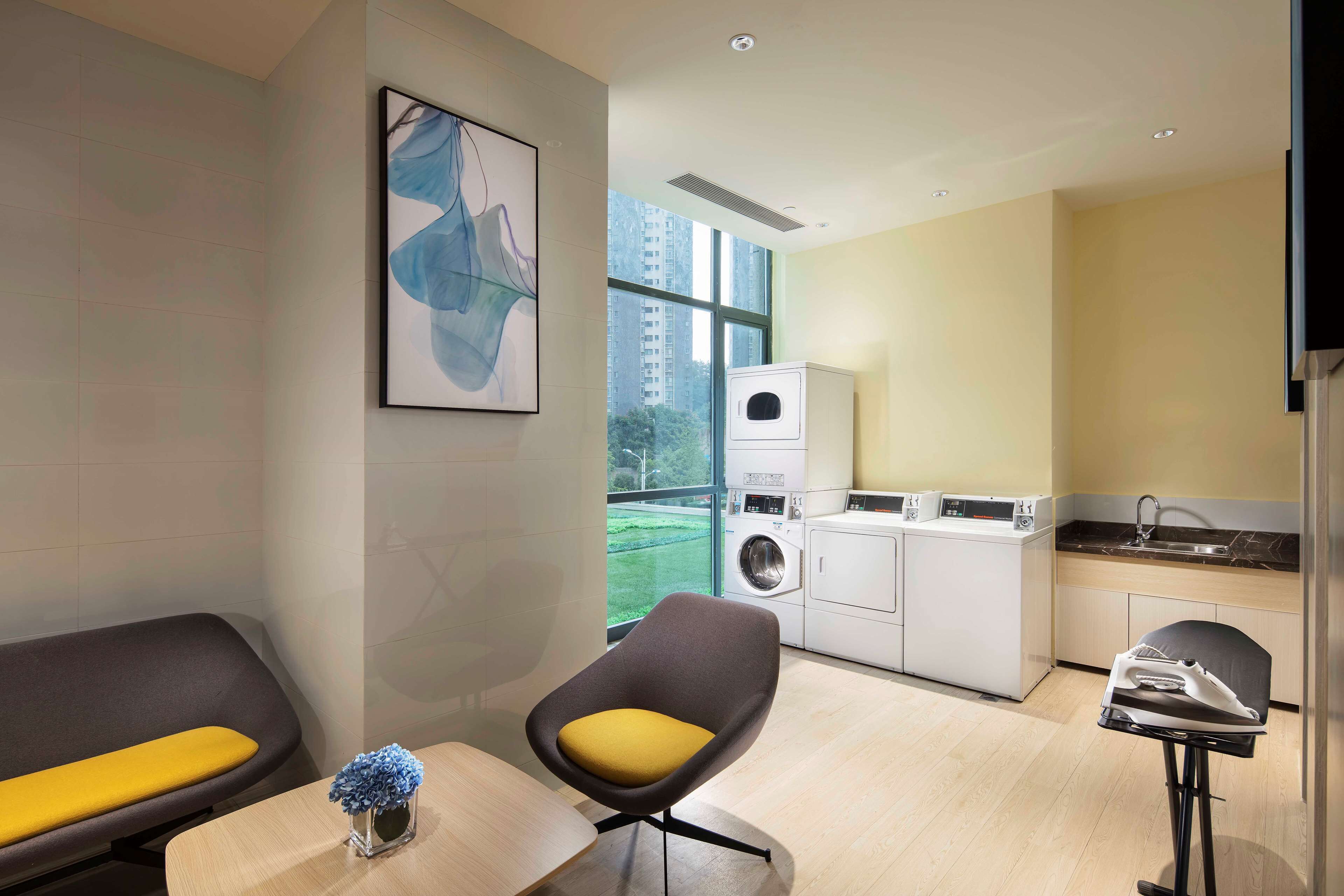 Una habitación de hotel moderna con sofá, una mesita con un florero, una silla, lavadora y una zona de cocina.