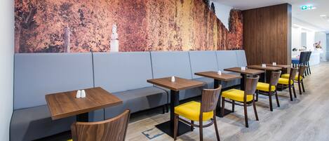 Un restaurante moderno con mesas de madera y sillas amarillas, un sofá gris y un mural grande en la pared.