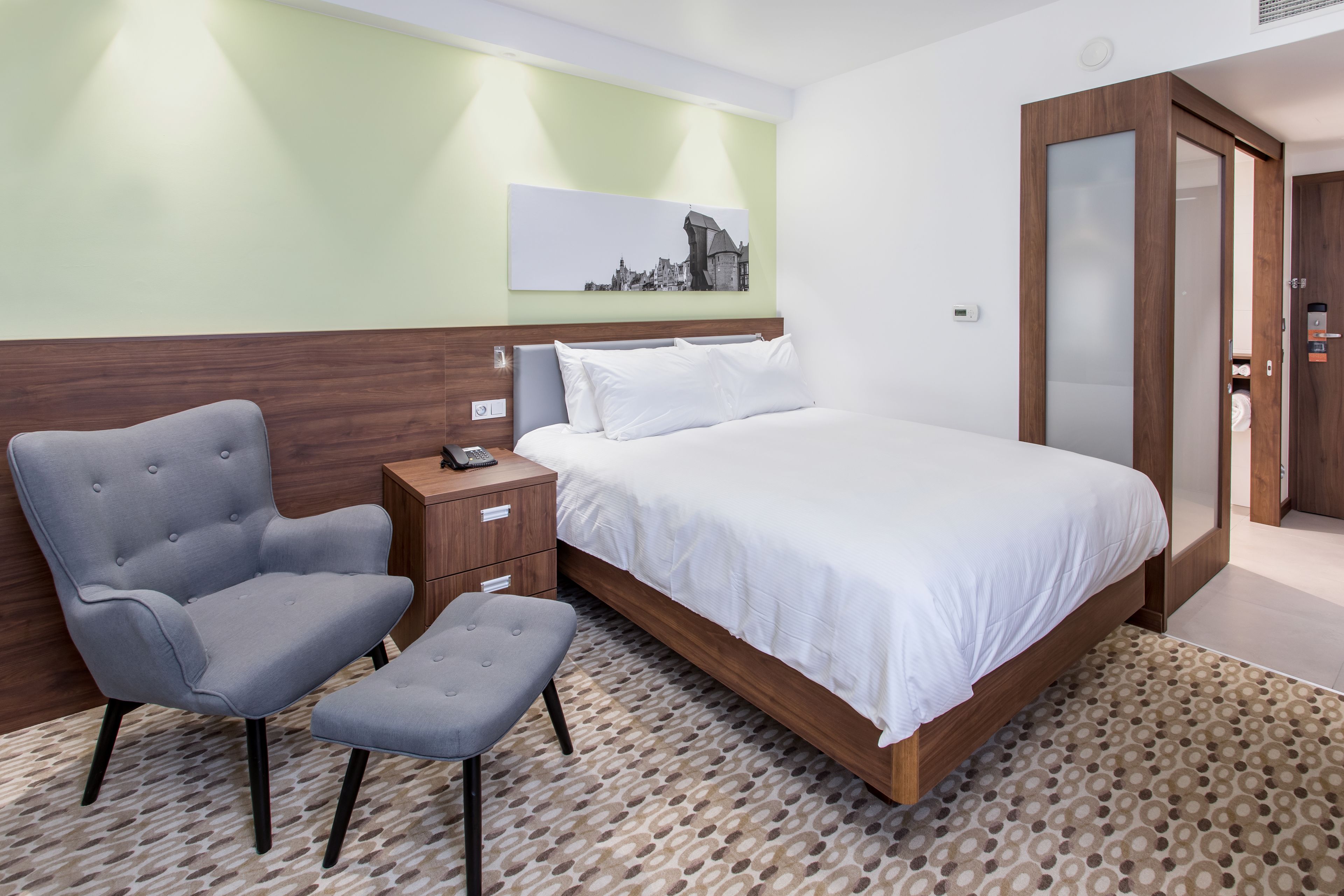 A modern hotel room with a bed, a grey armchair, a small table, and a patterned rug.