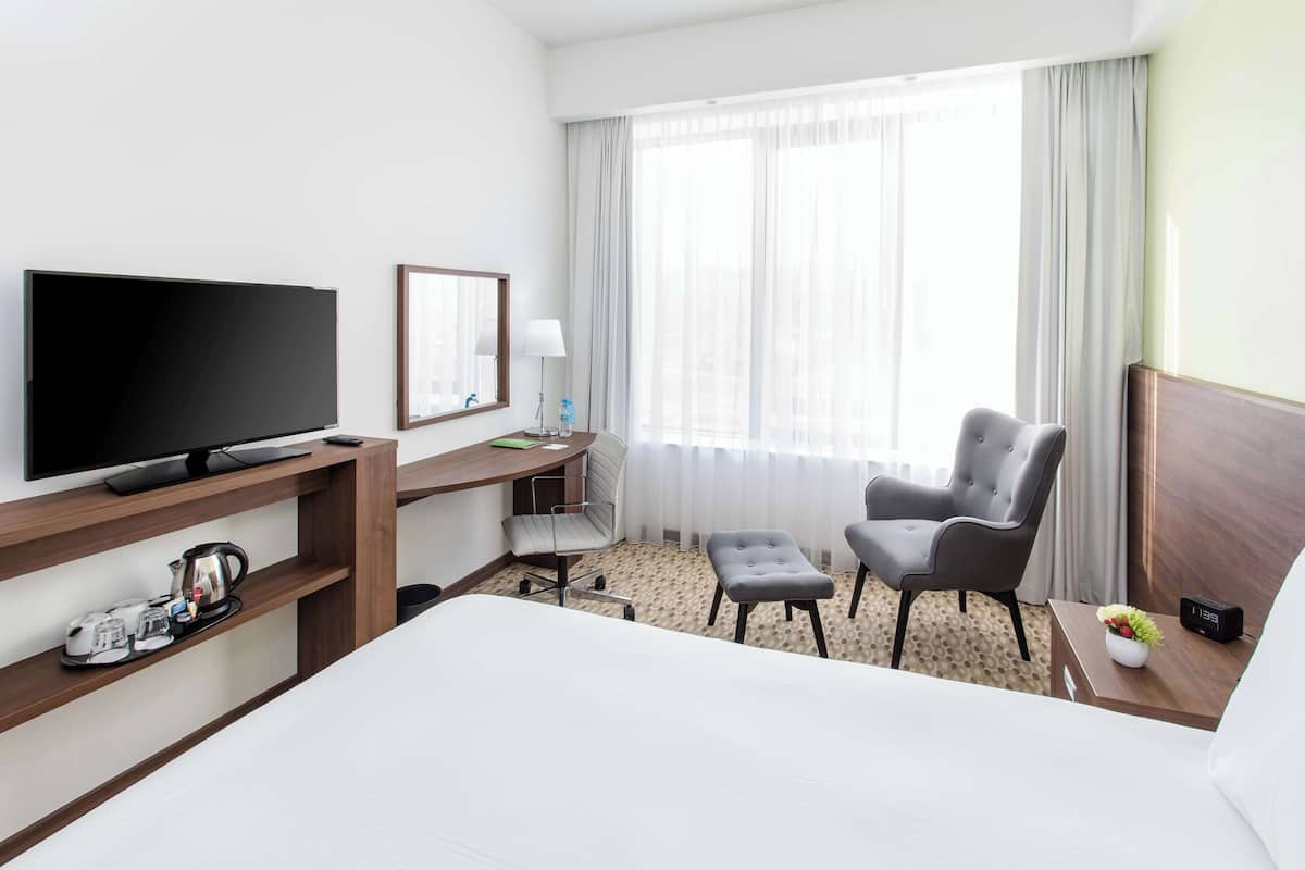 Een hotelkamer met een bed, een bureau met een televisie, een stoel, een klein tafeltje met een klok en een raam met gordijnen.