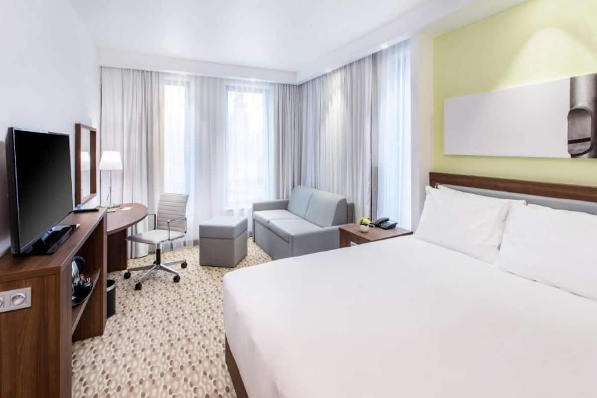Een hotelkamer met een bed, een bureau met een televisie, een stoel, een bank en een groot raam met gordijnen.