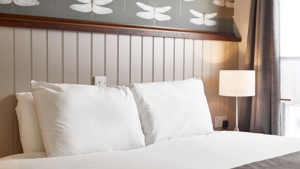 Un lit bien fait, avec du linge de lit blanc et un plaid gris, une tête de lit en bois à panneaux verticaux et une lampe de chevet.