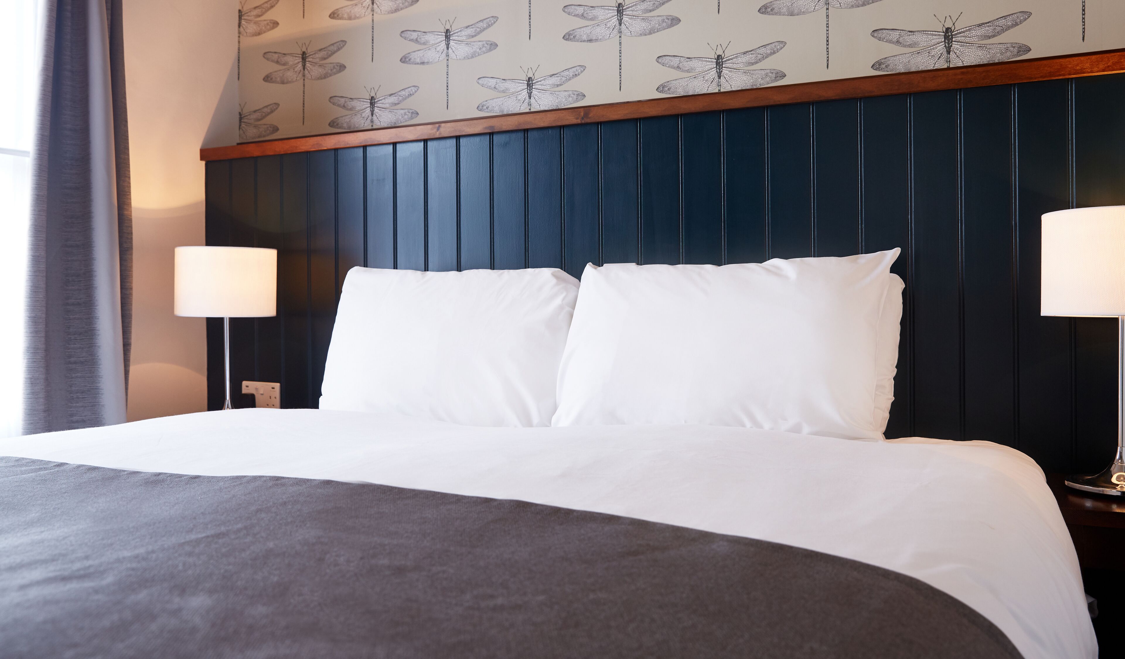 Ein Hotelzimmer mit einem Bett, weißer Bettwäsche, einem dunkelblauen Kopfteil und einer Wand mit Libellenmuster.