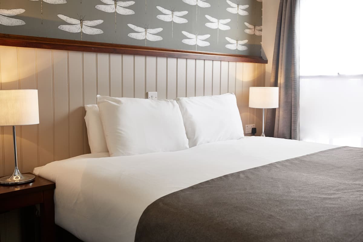Quarto de hotel com uma cama, duas luminárias de cabeceira, uma cabeceira de madeira e um papel de parede com padrão de borboletas.