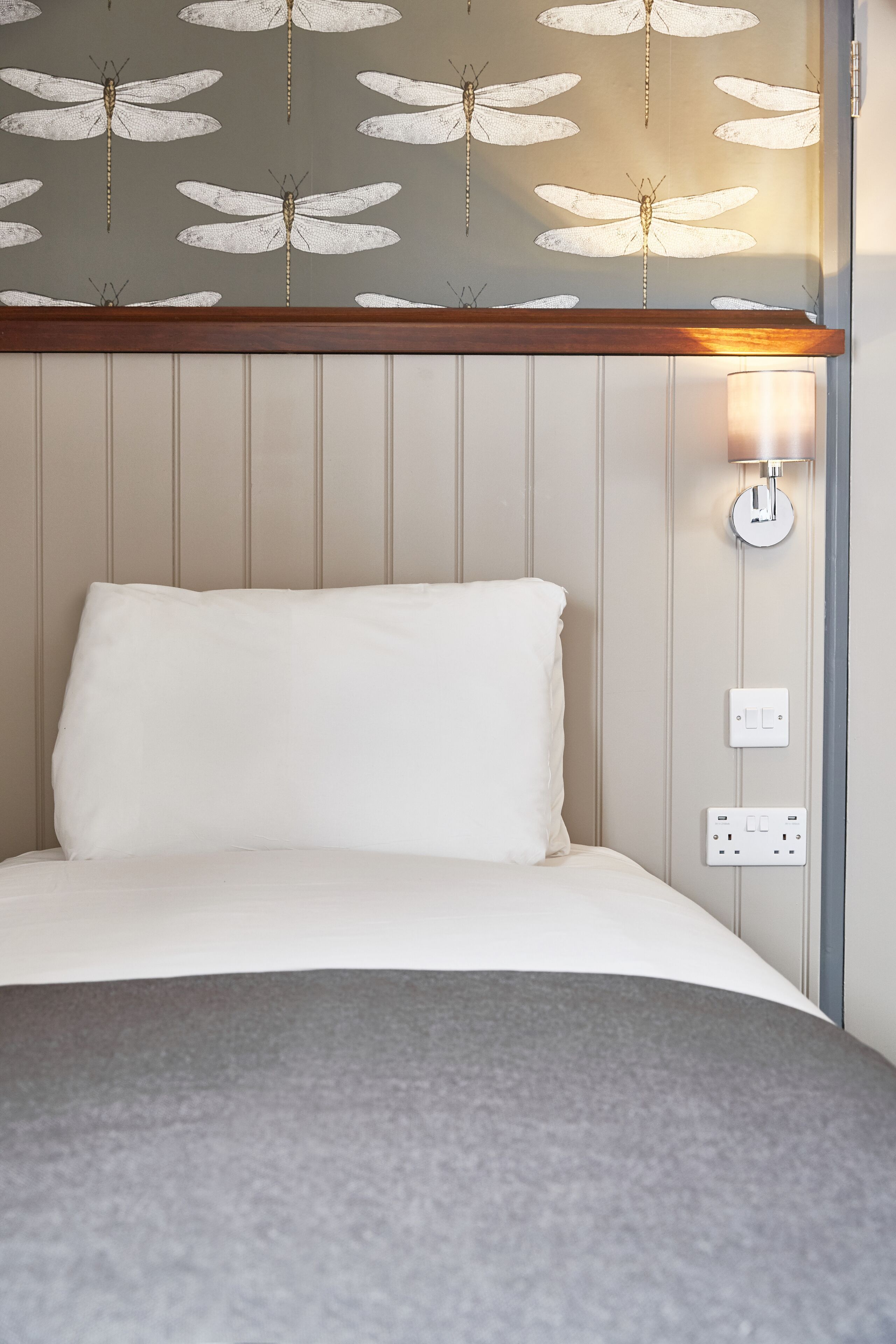 A hotel room with a bed, a wall-mounted light fixture, and a power outlet.