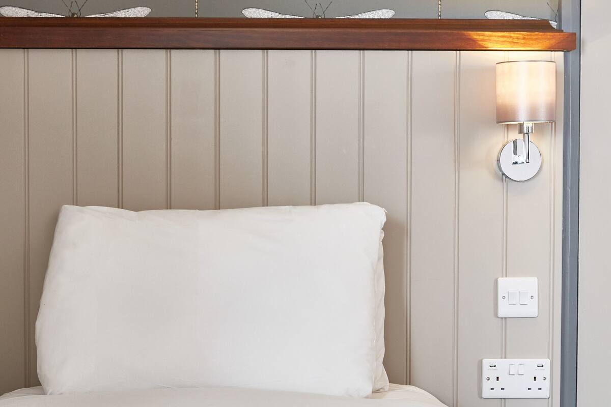 Quarto de hotel com cama, luminária fixada na parede e tomada.