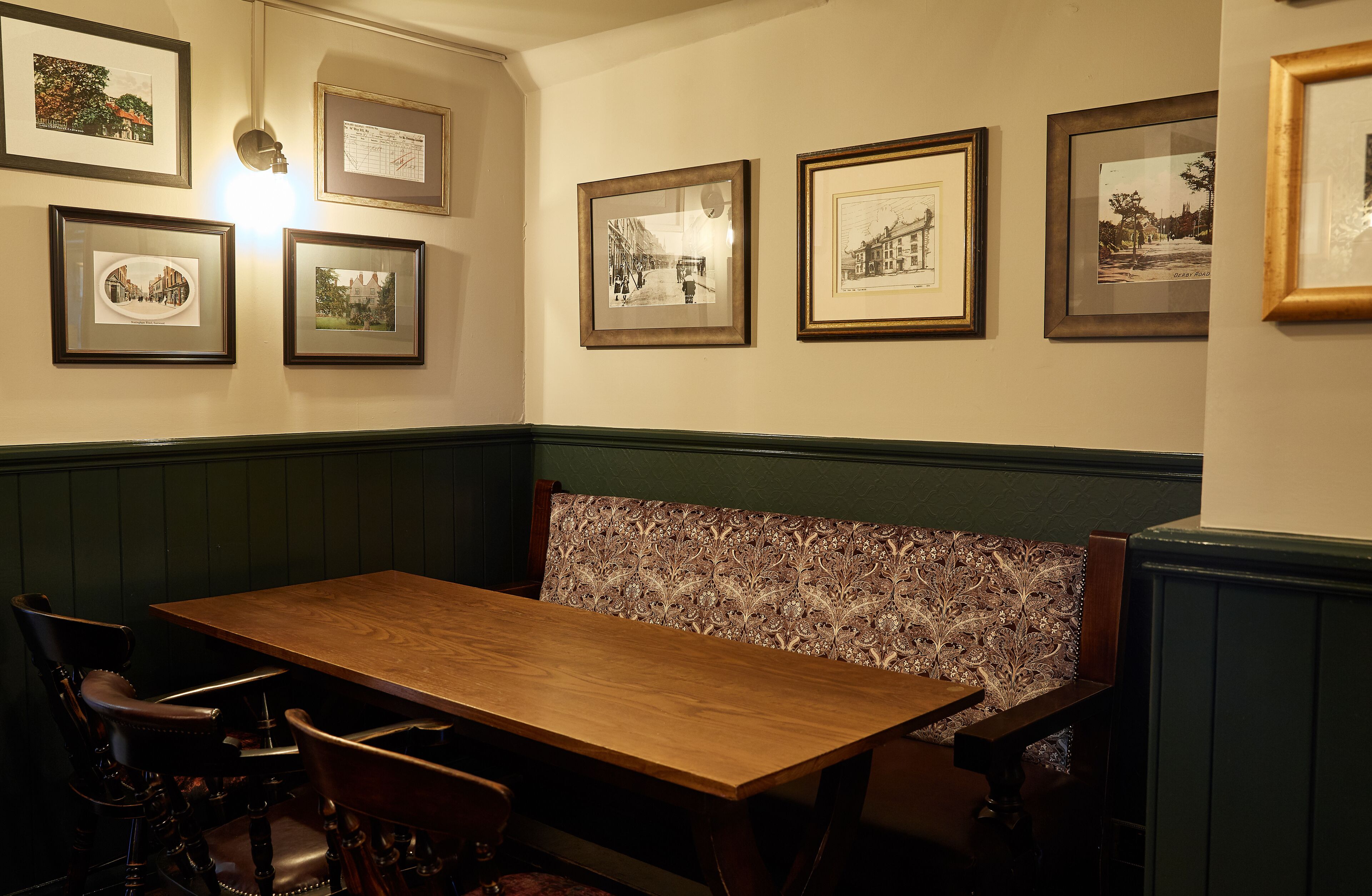 A dining area with a wooden table and upholstered bench, framed pictures on the wall, and green paneled walls.