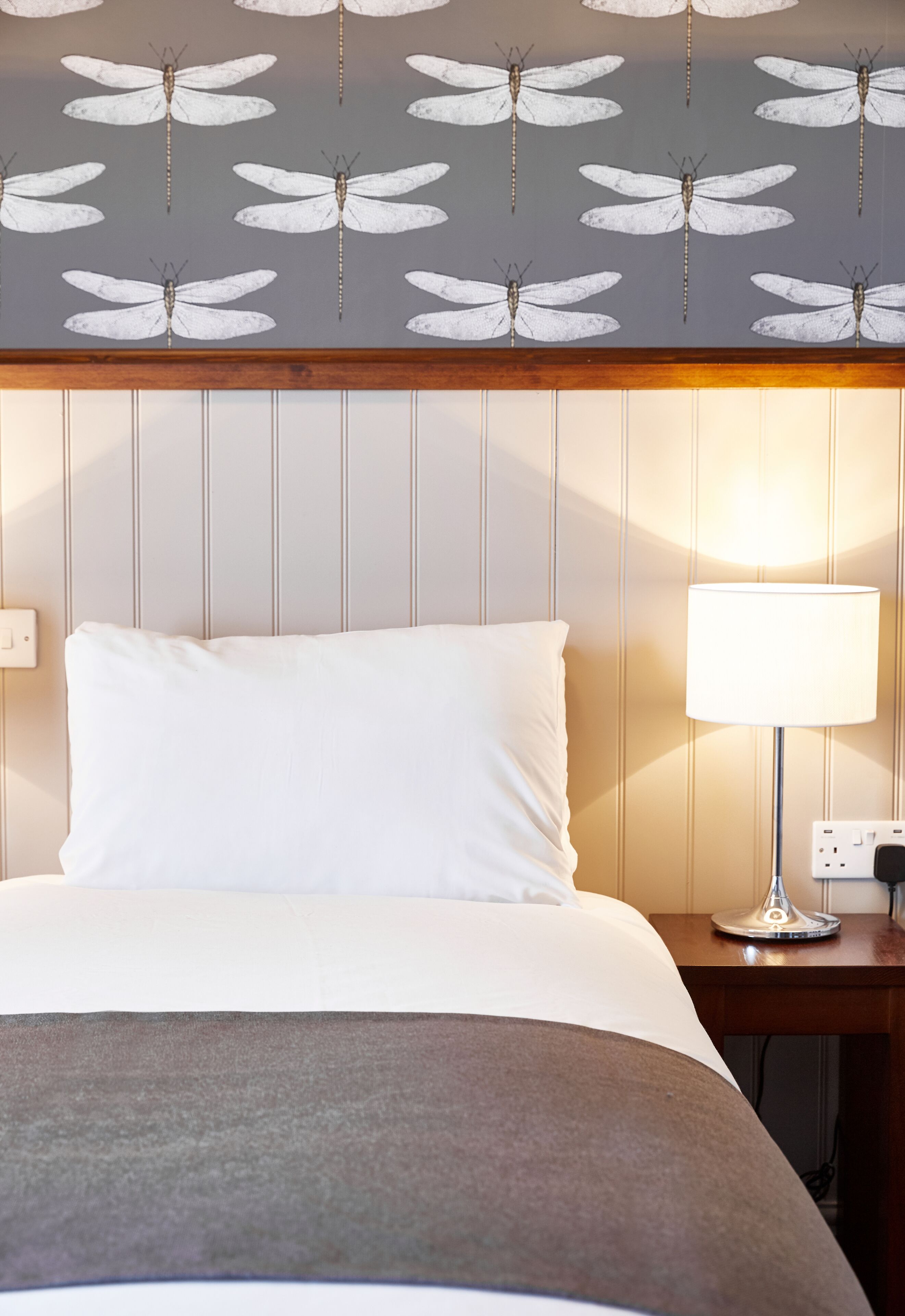 A neatly made bed with white linens, a bedside lamp, and a wall with a dragonfly pattern.