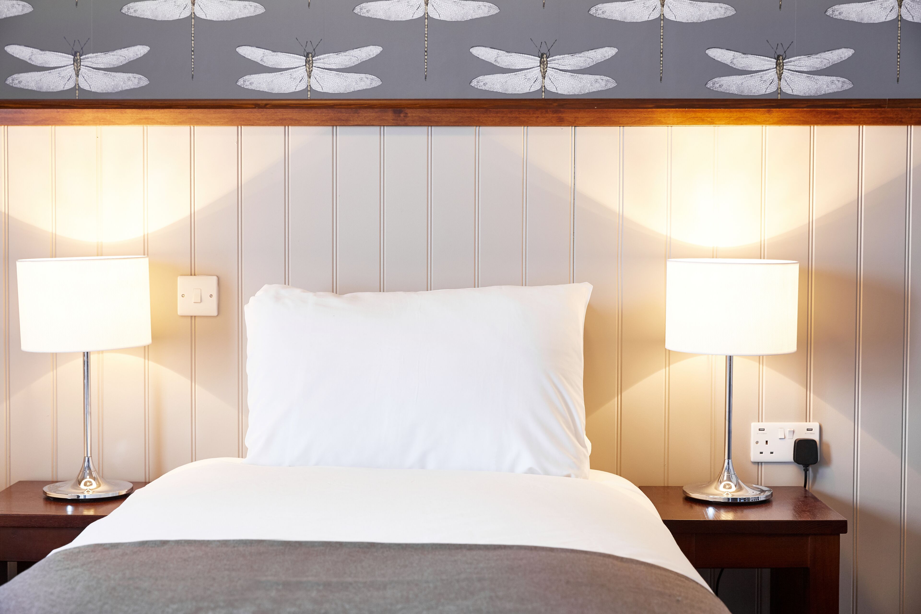 Une chambre d’hôtel avec un lit, deux lampes de chevet, une prise de courant et un mur orné d’un motif représentant des libellules.