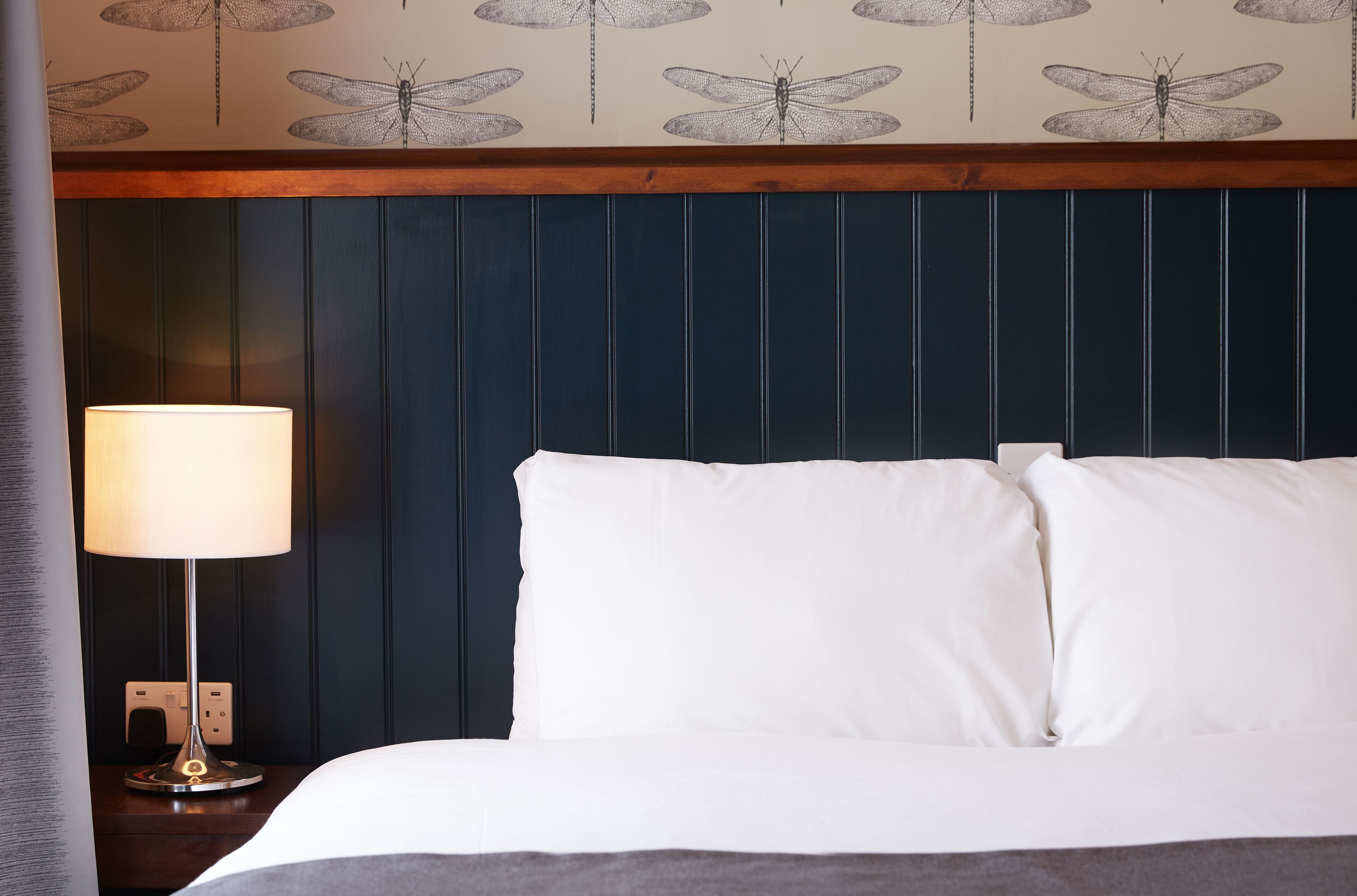 A neatly made bed with white linens, a bedside lamp, and a wall with a dragonfly pattern.