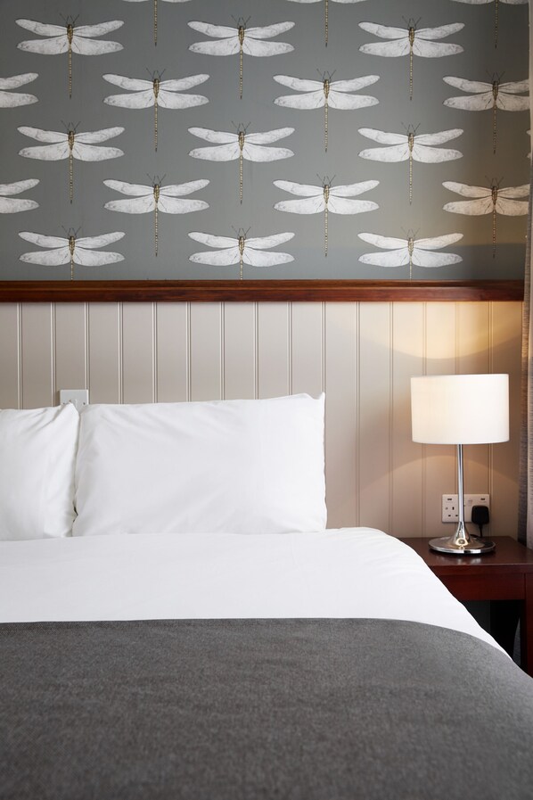 A hotel room with a bed, white bedding, a grey throw blanket, a nightstand with a lamp, and a wall with a dragonfly pattern.
