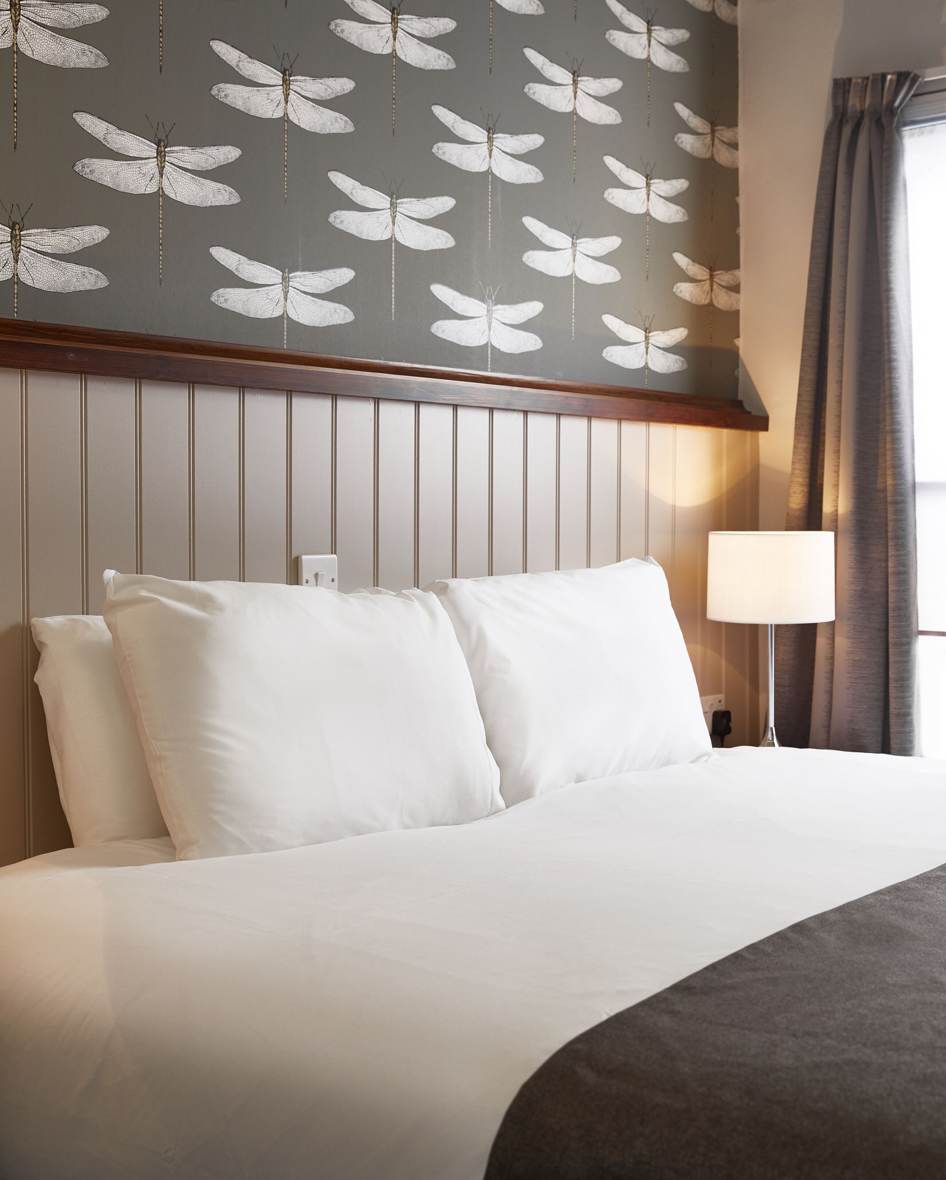 A hotel room with a bed, pillows, a nightstand, a lamp, and a wall with butterfly patterns.