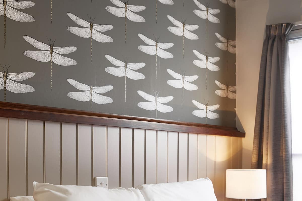 Quarto de hotel com cama, almofadas, mesa de cabeceira, abajur e parede com padrão de borboletas.