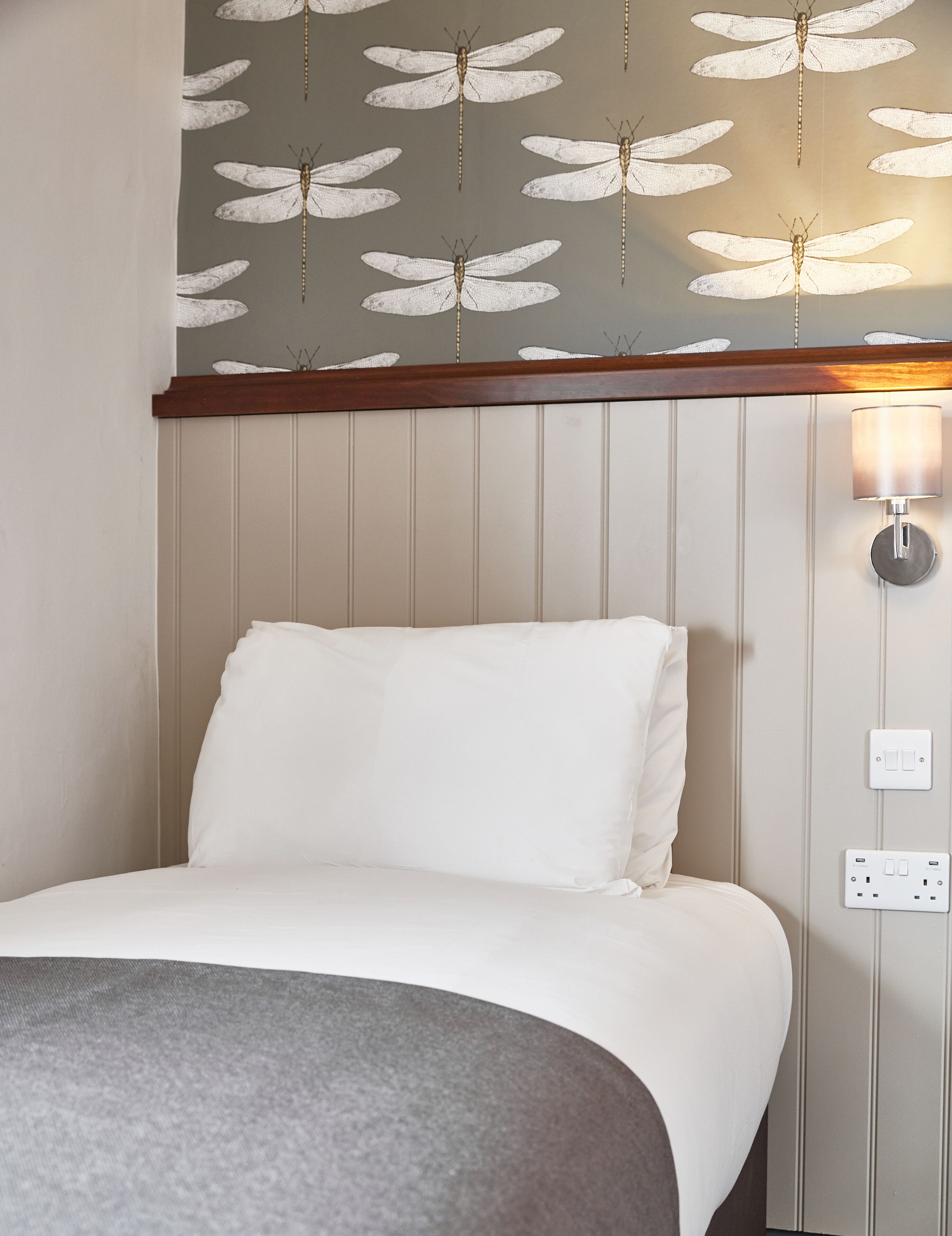 A hotel room with a bed, white bedding, a grey throw, a wall with a dragonfly pattern, a wooden headboard, and a wall-mounted light fixture.