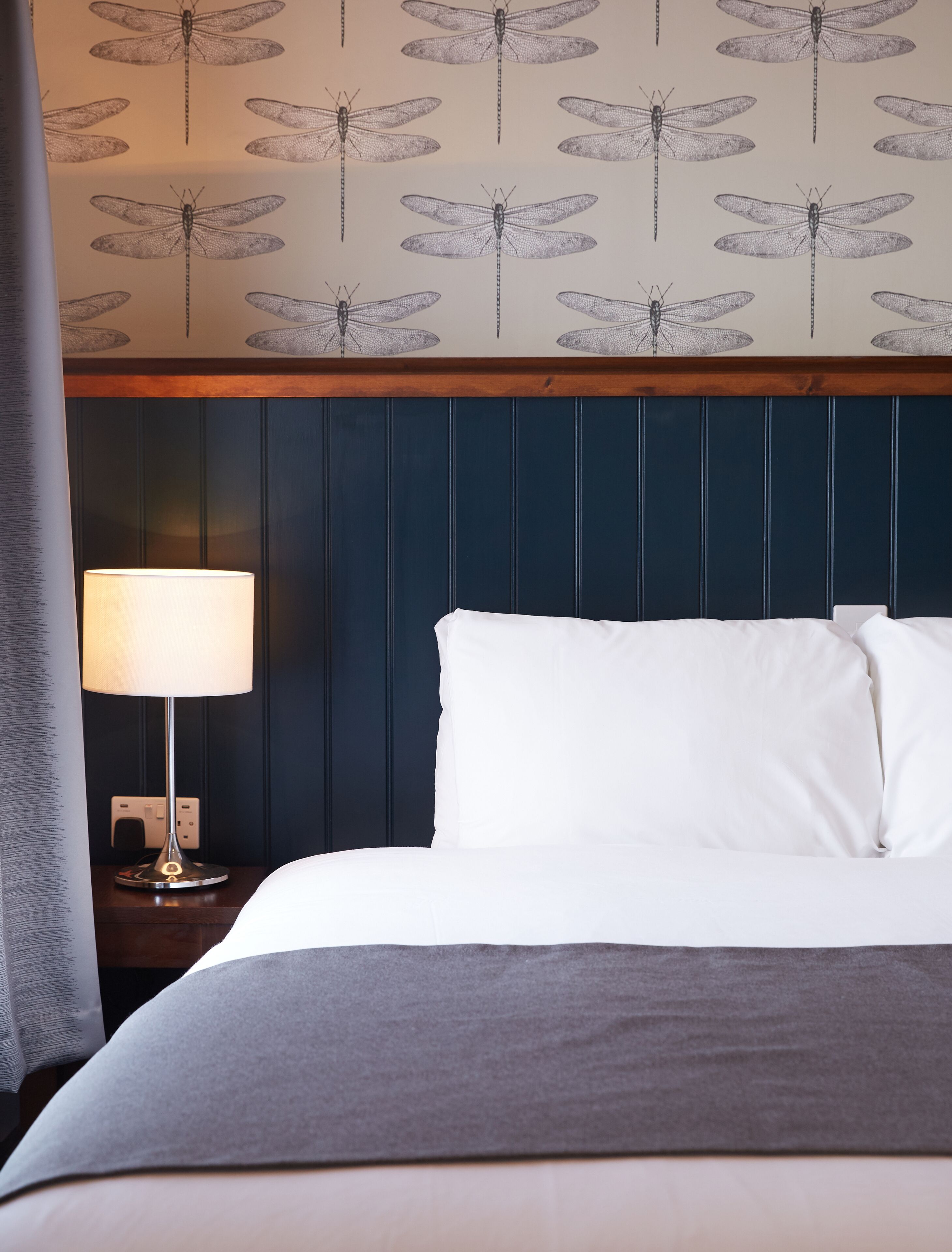 A hotel room with a bed, a nightstand with a lamp, and a wall with a dragonfly pattern.