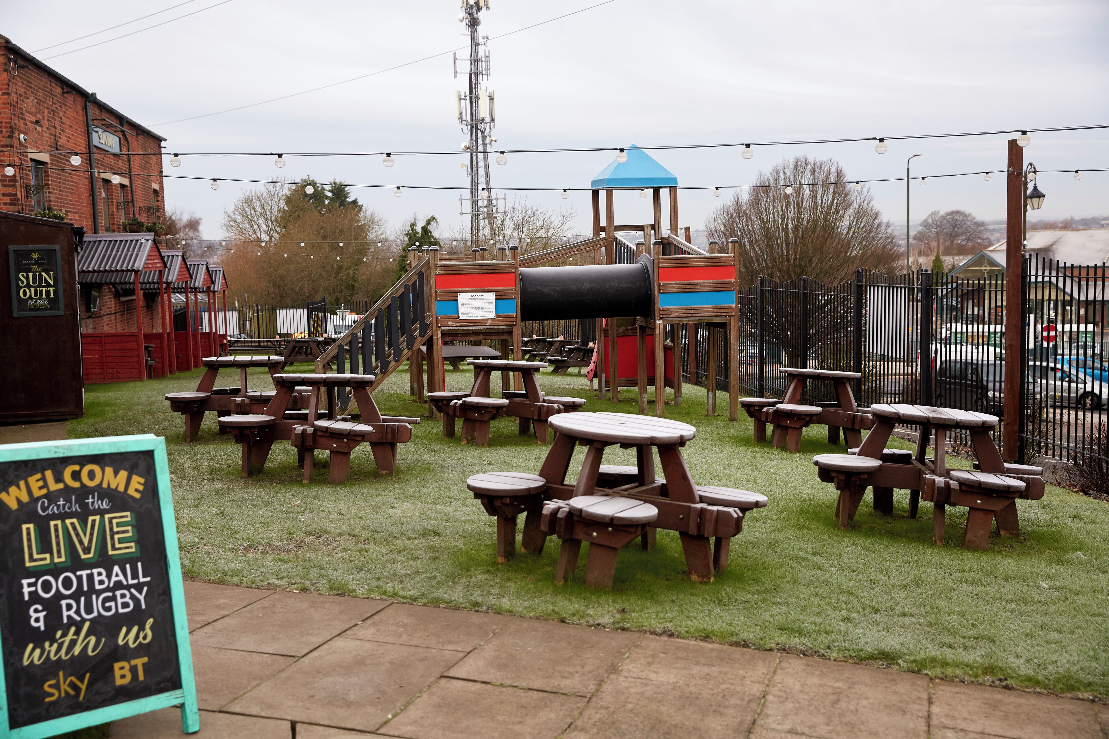 An outdoor seating area with picnic tables and a play structure, surrounded by a fence and a building with a sign.